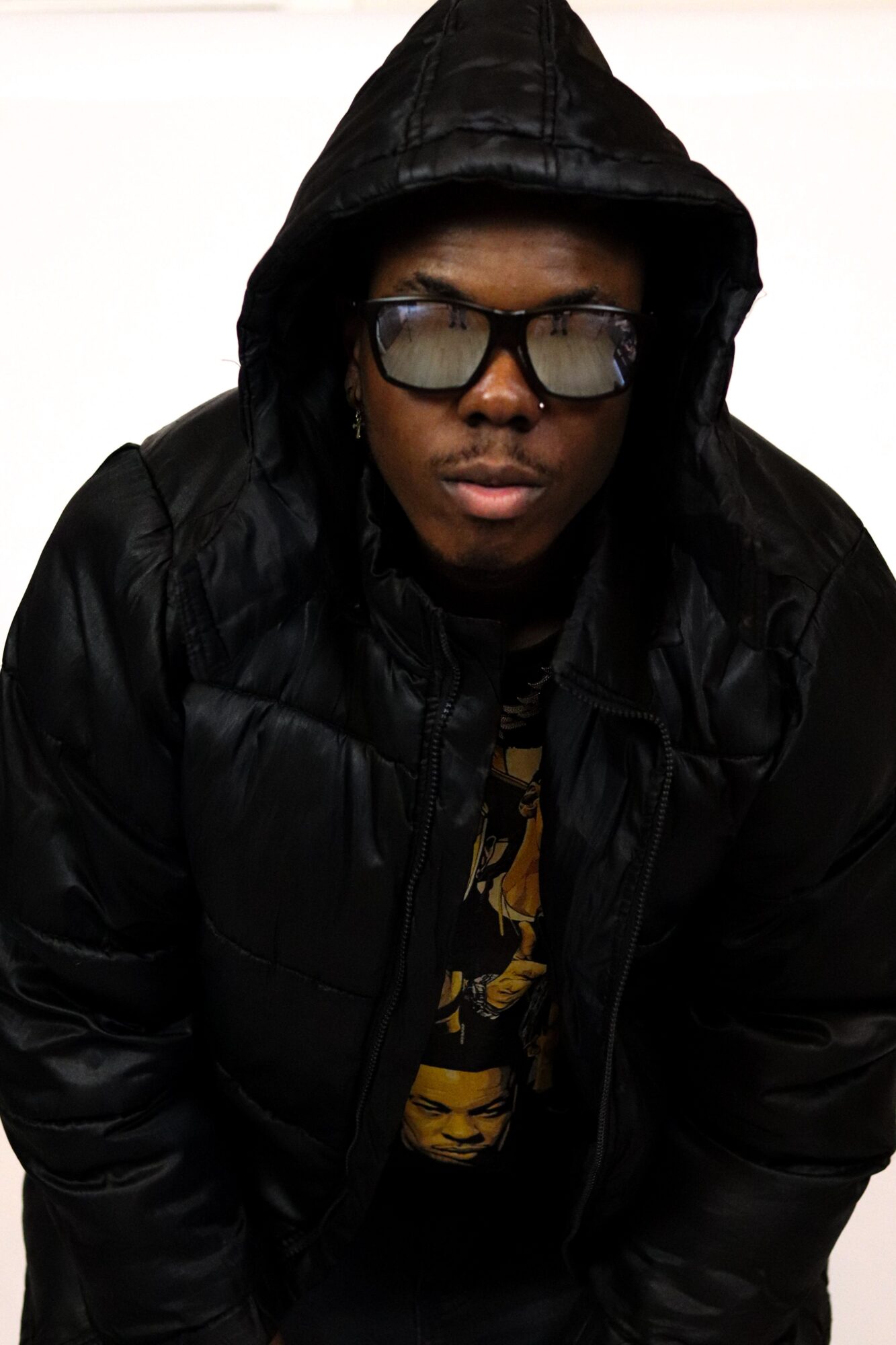 Person wearing a black hooded jacket and glasses, looking down, against a plain background.