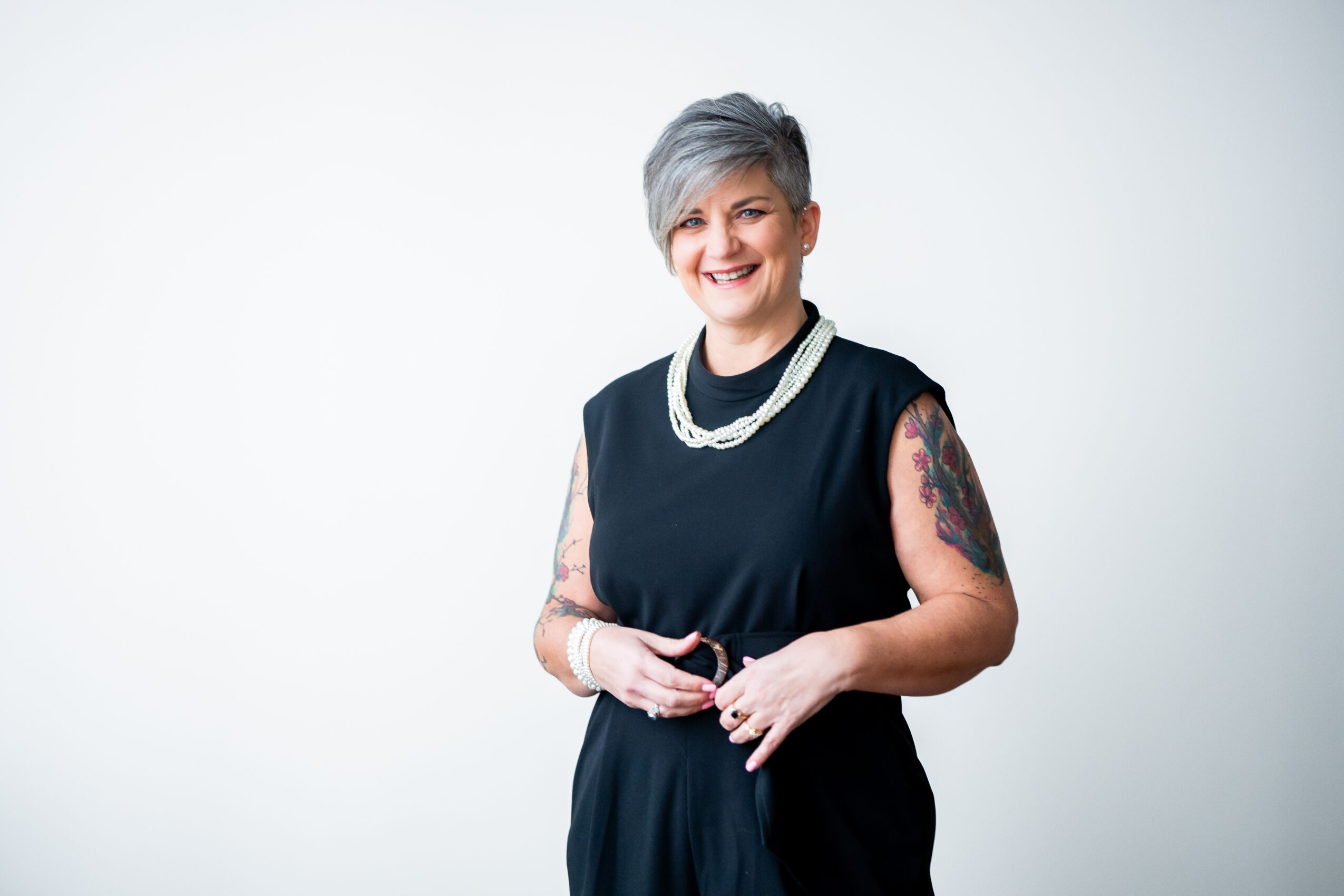 Smiling woman with short gray hair, wearing a black sleeveless dress and pearl necklace, standing against a white background.