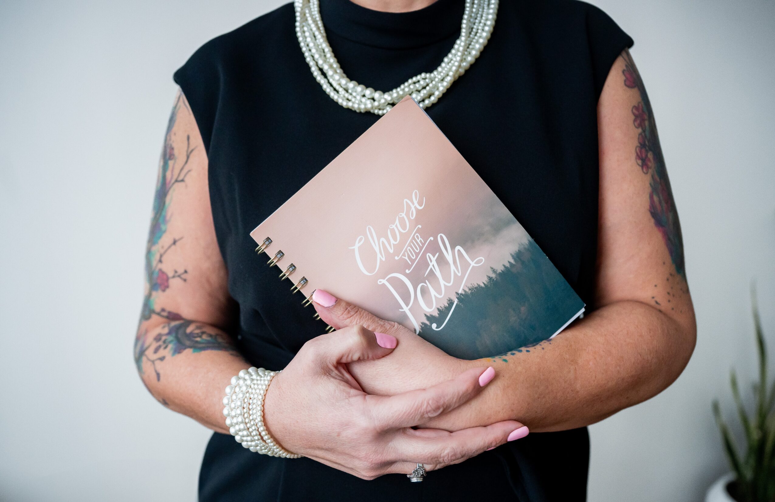 Person holding a notebook titled 'Change of Path' with tattoos, pearl jewelry, and a black sleeveless top.