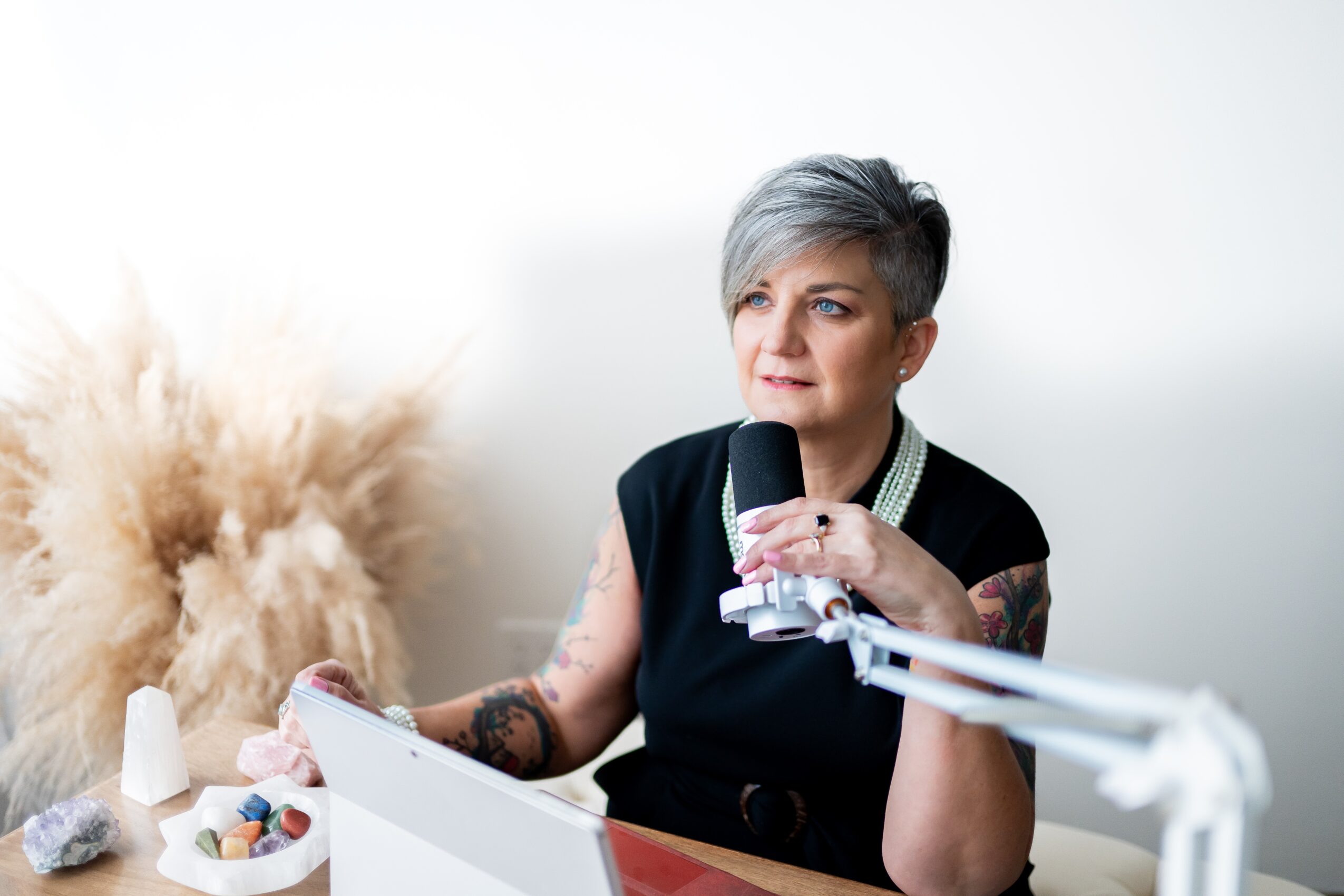 Woman with short gray hair holding a microphone, sitting at a desk with a laptop, in a bright room with a fluffy plant.