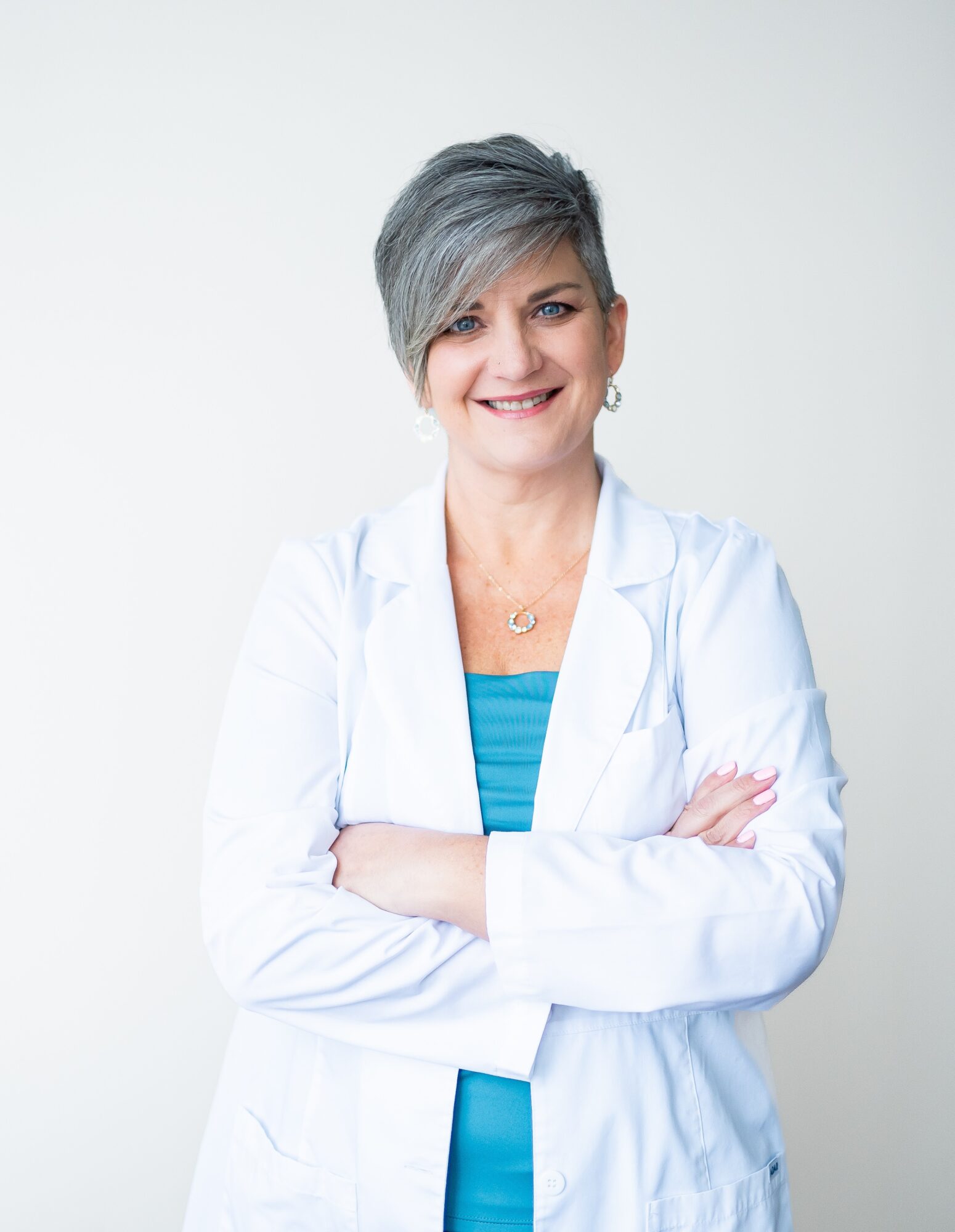 A smiling woman with short gray hair wearing a white lab coat and blue top, arms crossed, against a plain background.