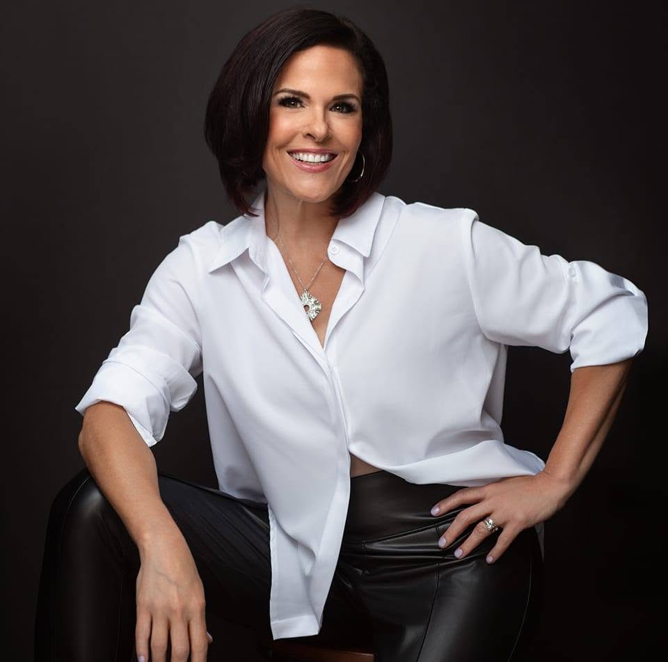 Woman with dark hair smiling, wearing a white shirt and black pants, sitting against a dark background.