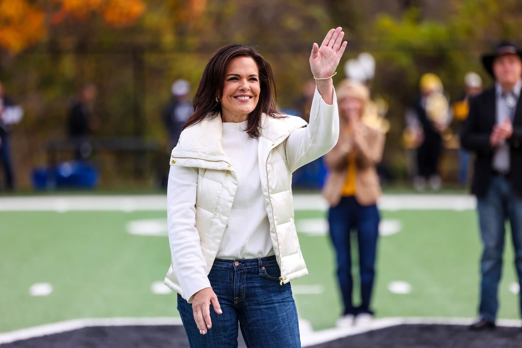 Woman smiling and waving outdoors on a sports field, with people in the background, trees, and a fence.