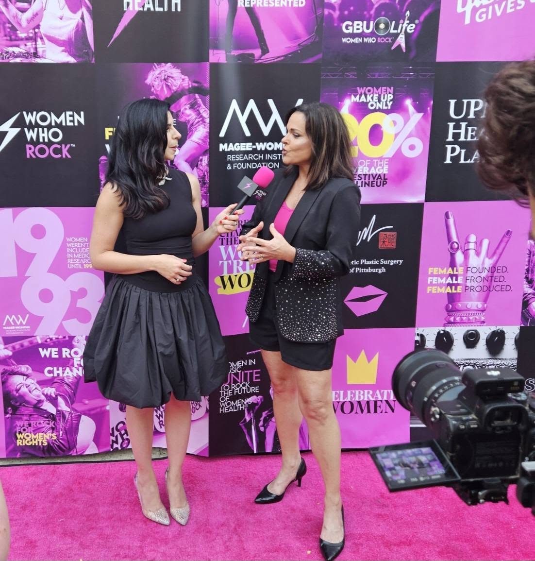 Two women are talking in front of a pink and black backdrop with various logos and text, one holding a microphone.