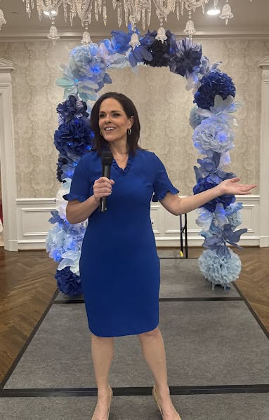 Woman in blue dress standing in front of a blue and white floral arch, smiling, holding a microphone, indoors.