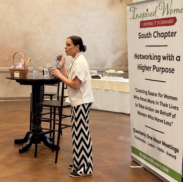 Woman speaking into microphone at a table with baskets and glasses, next to a large vertical banner, in a room with plain walls.