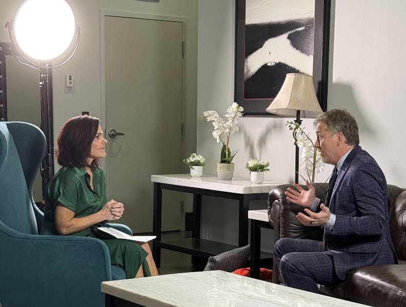 Woman in green dress and man in suit sit across from each other in a room, engaged in conversation, with a lamp and artwork behind them.