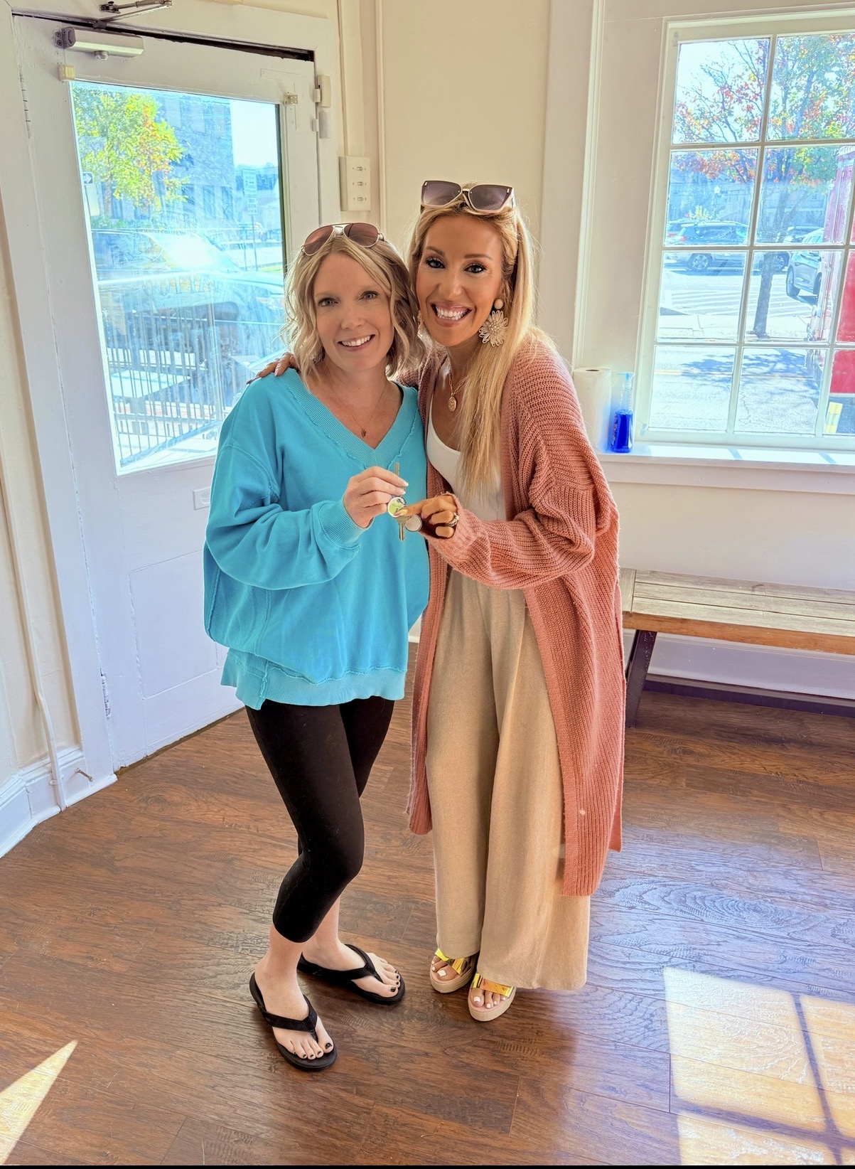 Two women smiling indoors, one in a blue top and black pants, the other in a long beige dress and pink cardigan, holding a small object, near a window with sunlight.