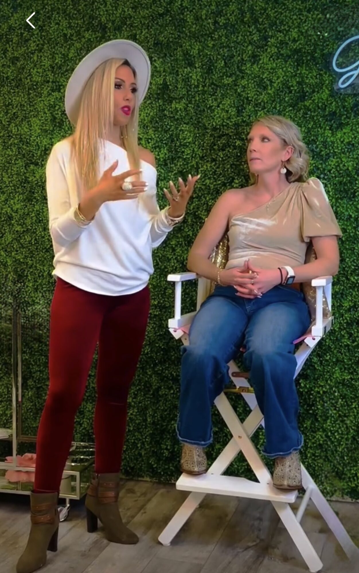Two women converse indoors, one standing with a hat and casual outfit, the other sitting on a white chair with a green background.