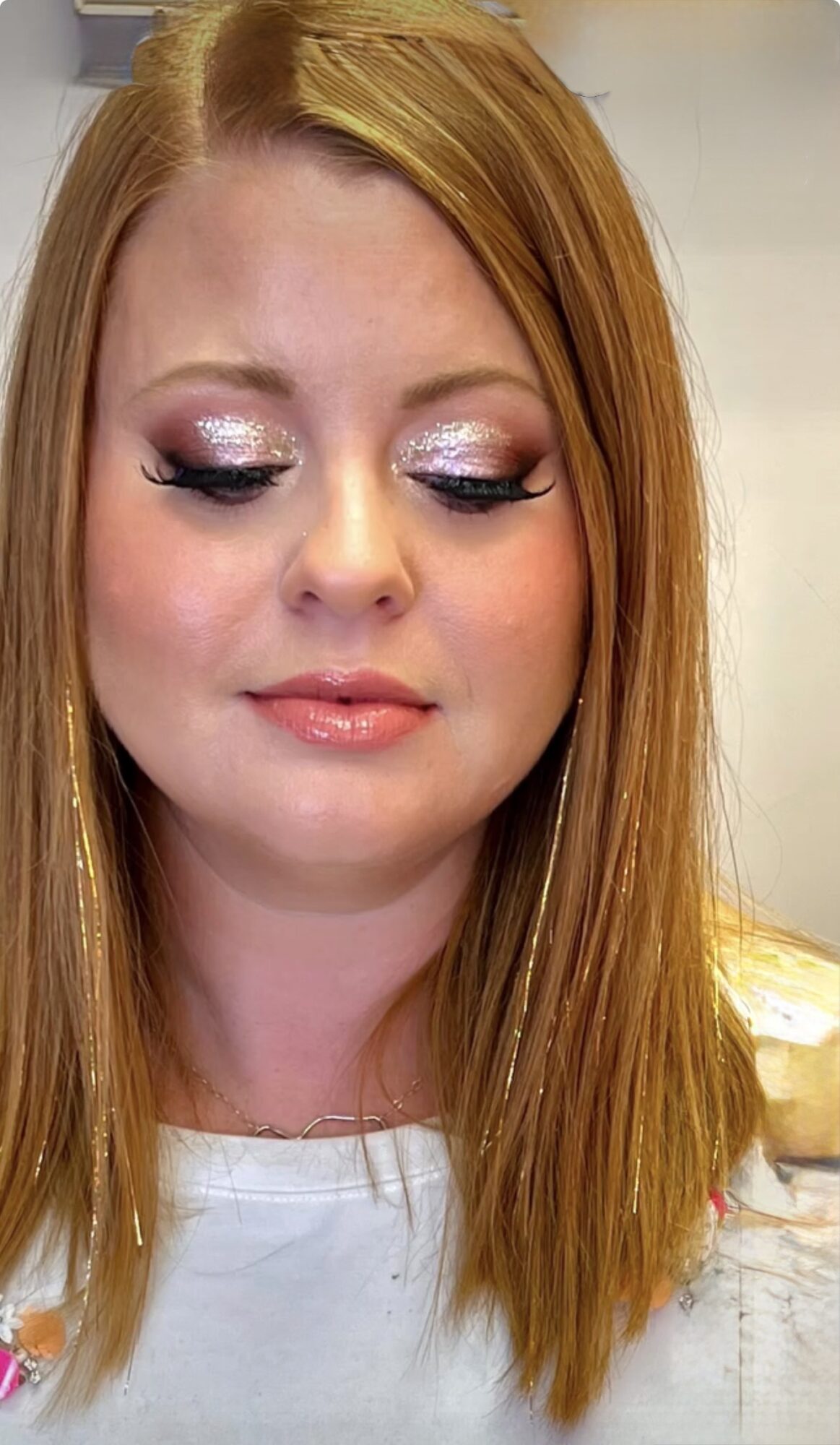 Close-up of a woman with red hair and makeup, looking downward, with a neutral background.
