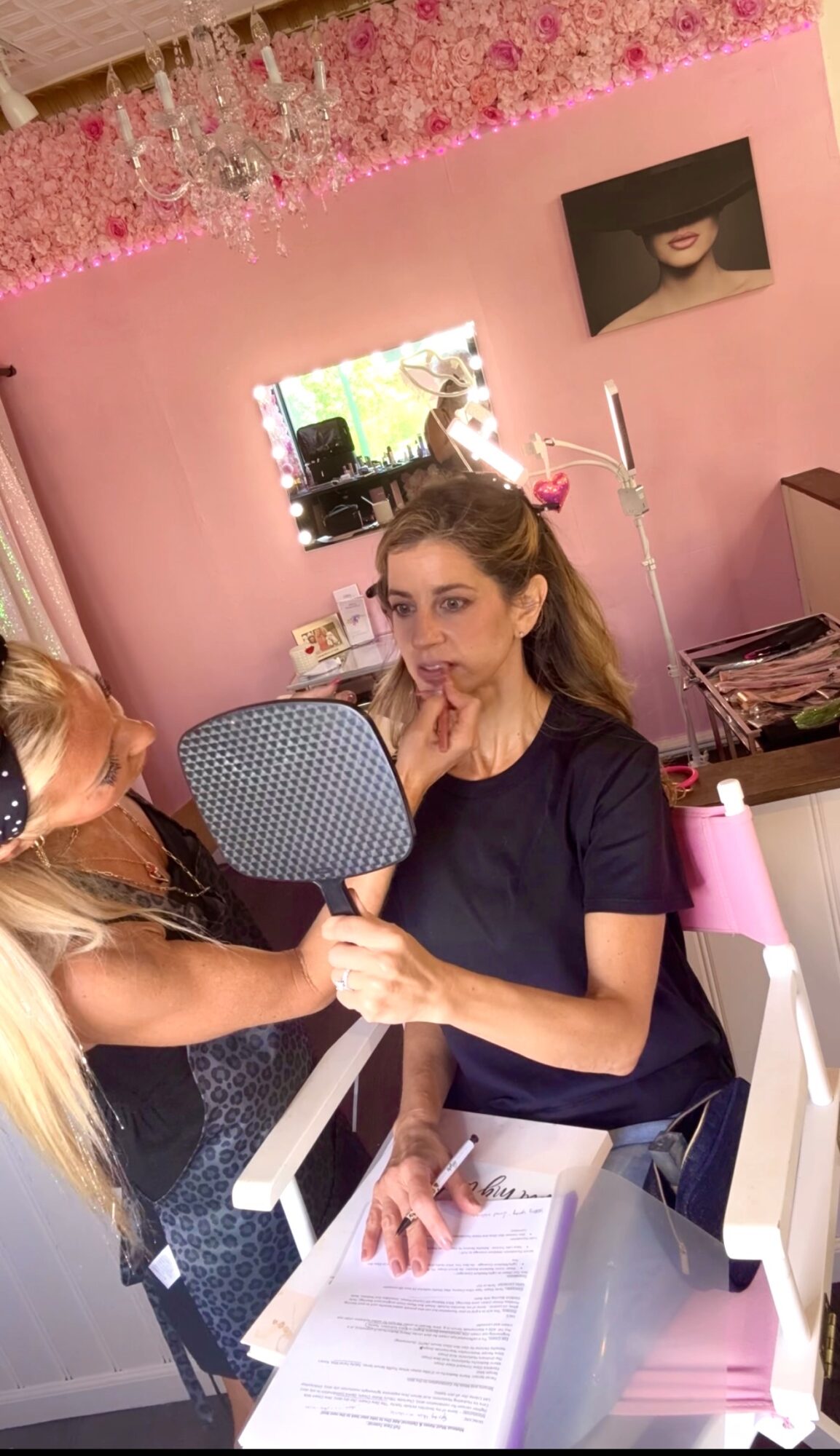Woman with makeup mirror and makeup artist in a pink room with a chandelier, TV, and framed photo on wall.
