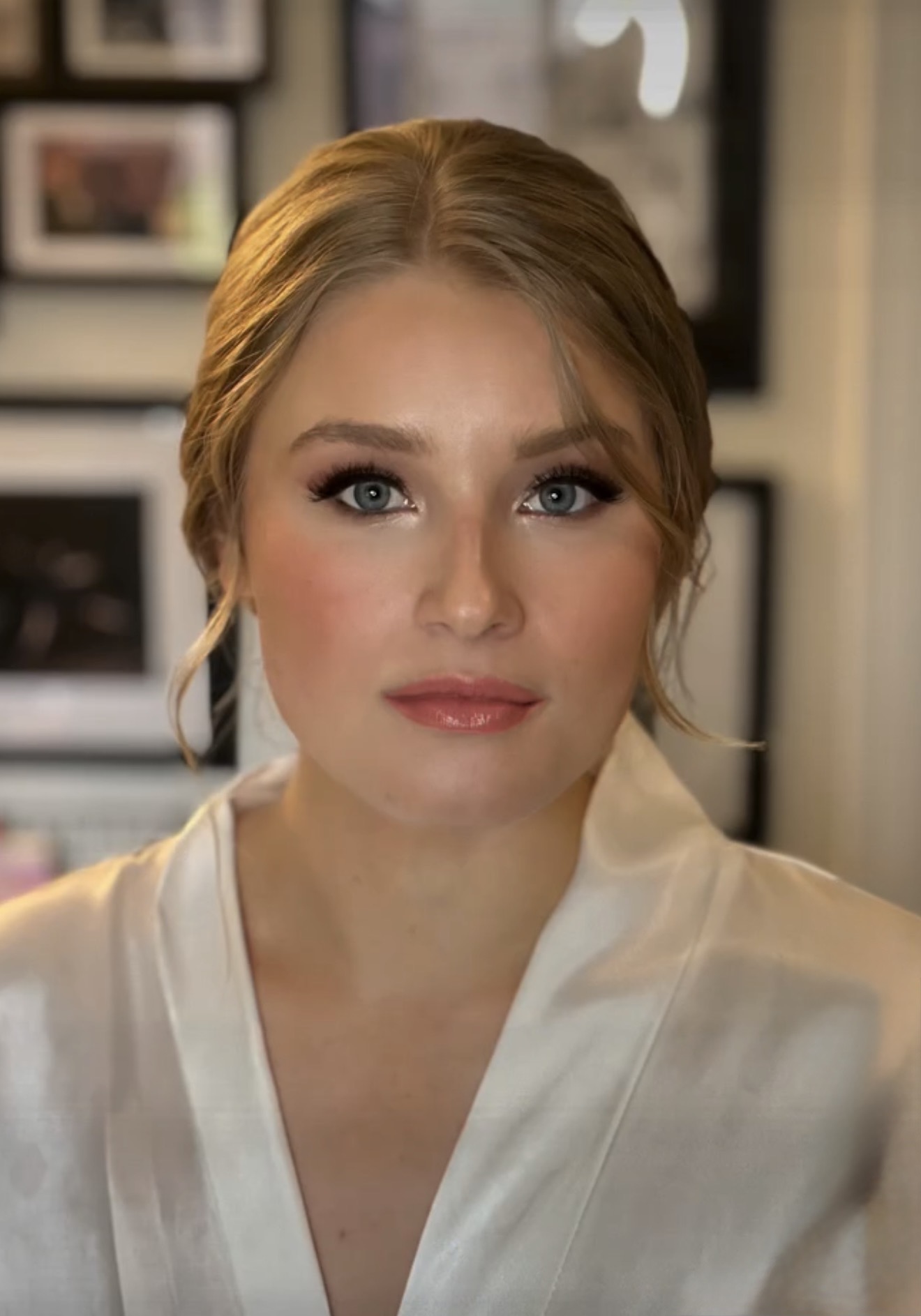 Portrait of a woman with blonde hair, blue eyes, and makeup, wearing a white top, indoors with framed pictures in the background.