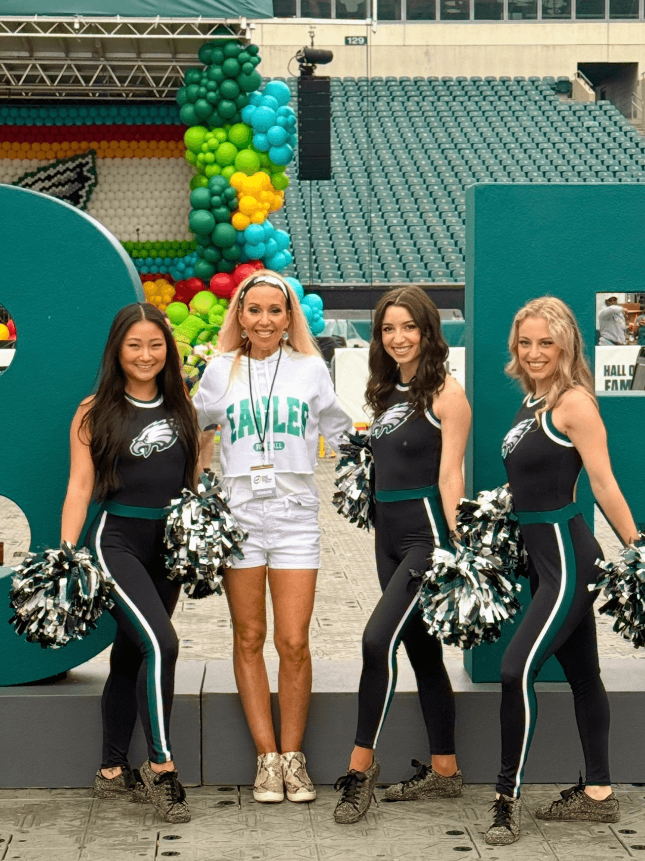 Four women, including a cheerleader, standing together at a stadium with balloons in the background.