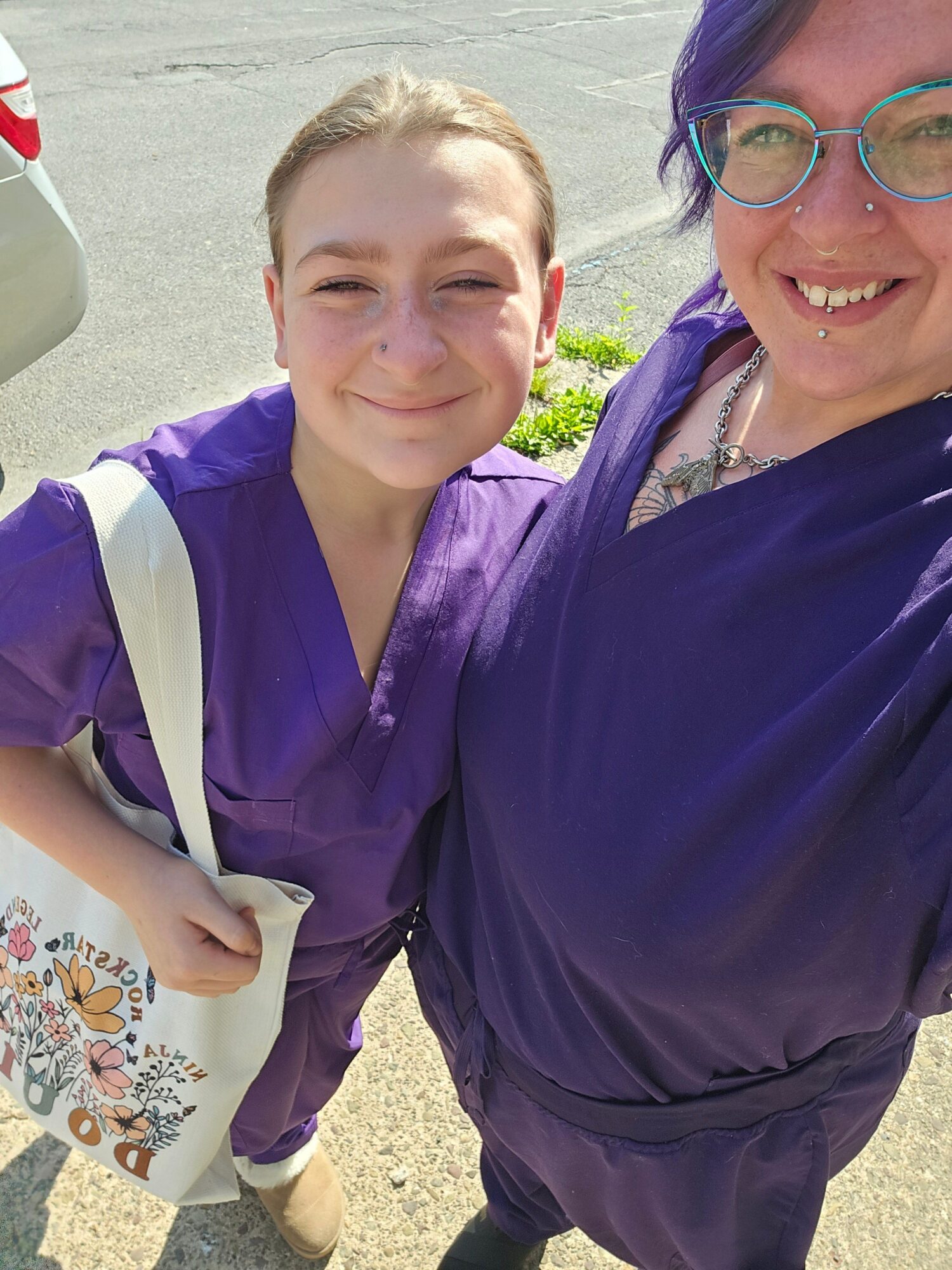 Two people smiling outdoors, one holding a paper with colorful drawings, the other wearing glasses and a purple jacket.