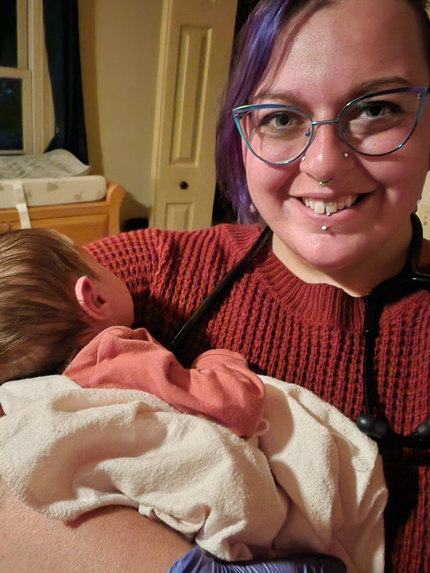 Person with glasses smiling, holding a baby wrapped in a blanket, in a room with a door and furniture in the background.