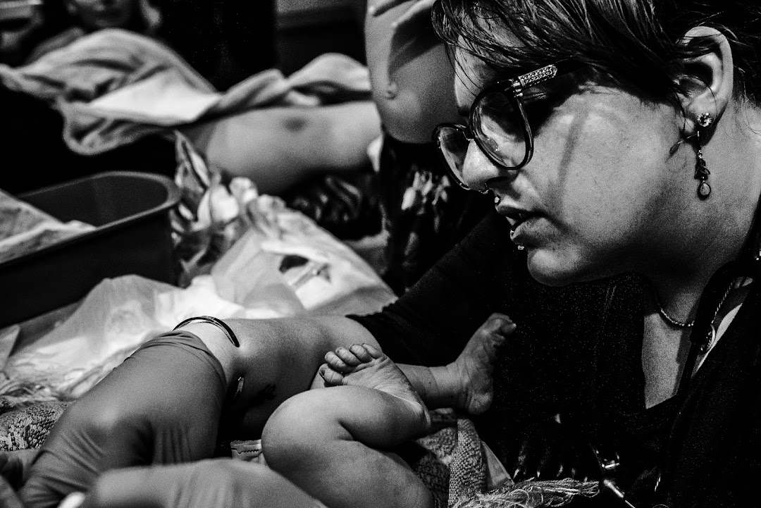 A woman with glasses and earrings looks at a baby lying on a surface, surrounded by various items.