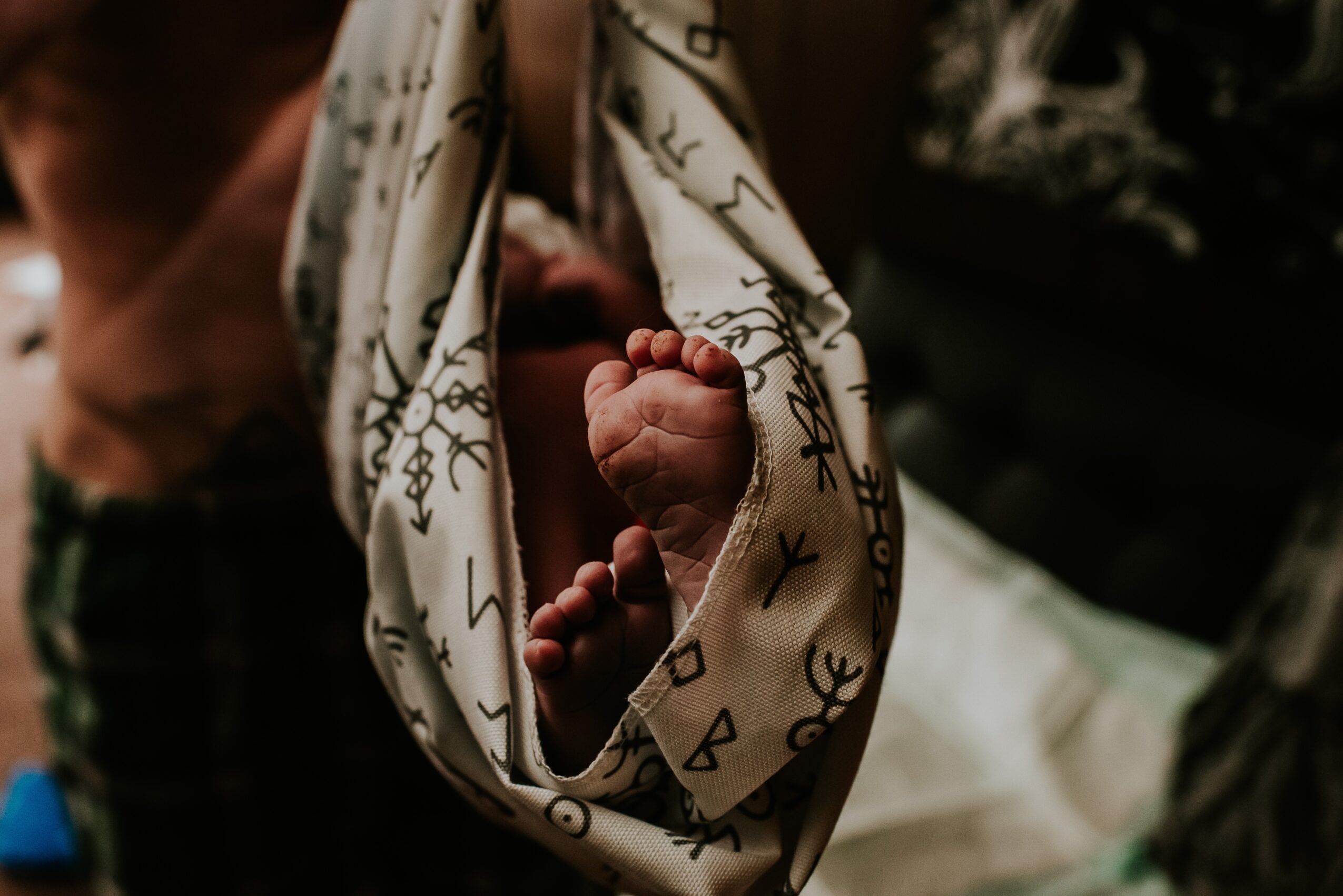Close-up of a newborn's feet wrapped in a patterned cloth, with a blurred person in the background.