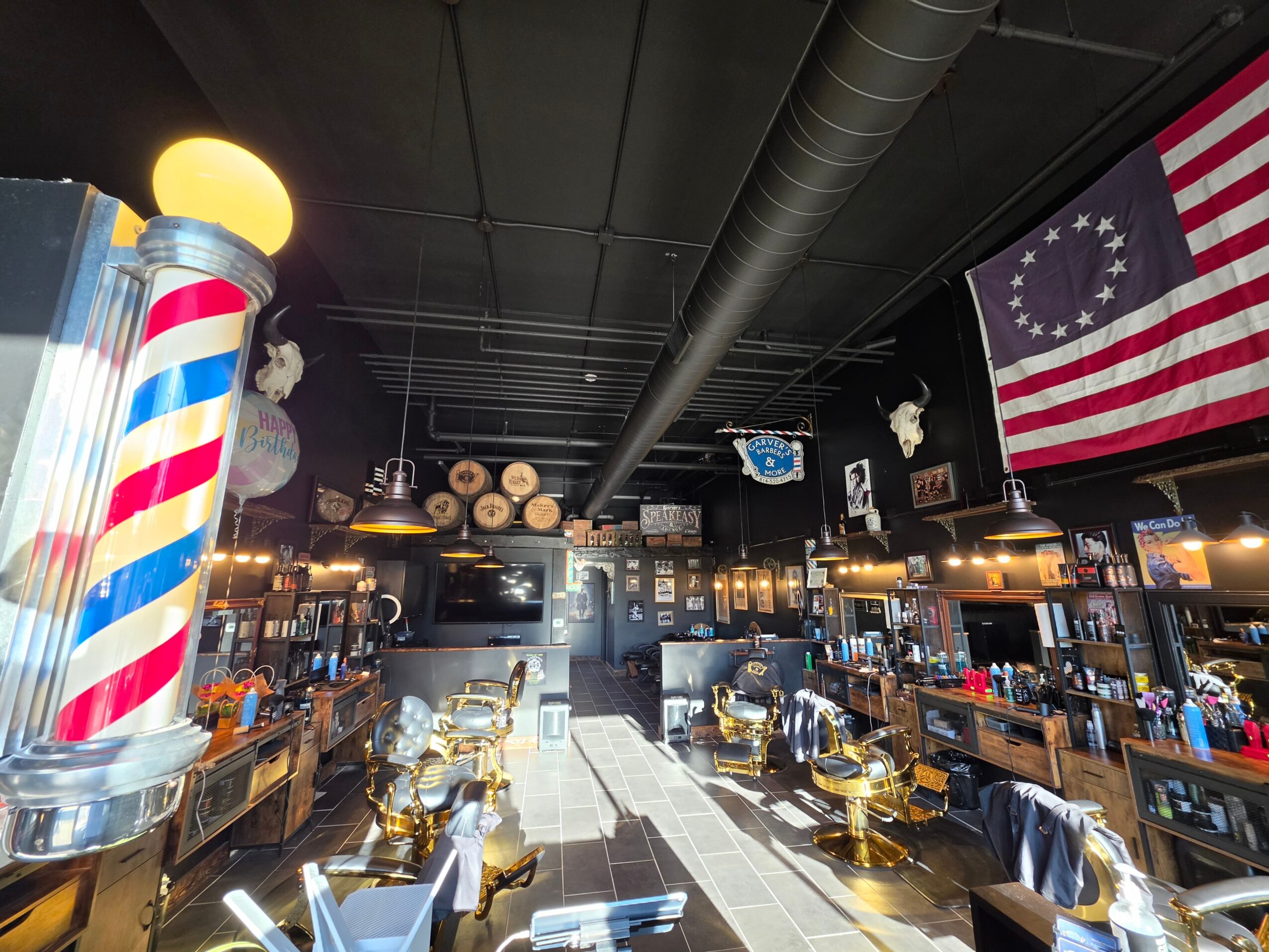 Bar interior with barber pole, American flag, and bar stools, illuminated by warm lighting, with ceiling pipes visible.