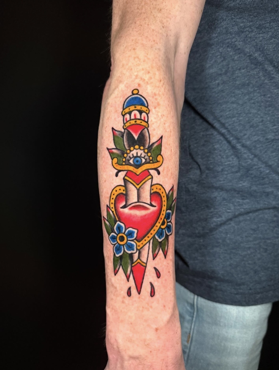 Colorful tattoo of a heart with flowers, a dagger, and decorative elements on person's forearm.