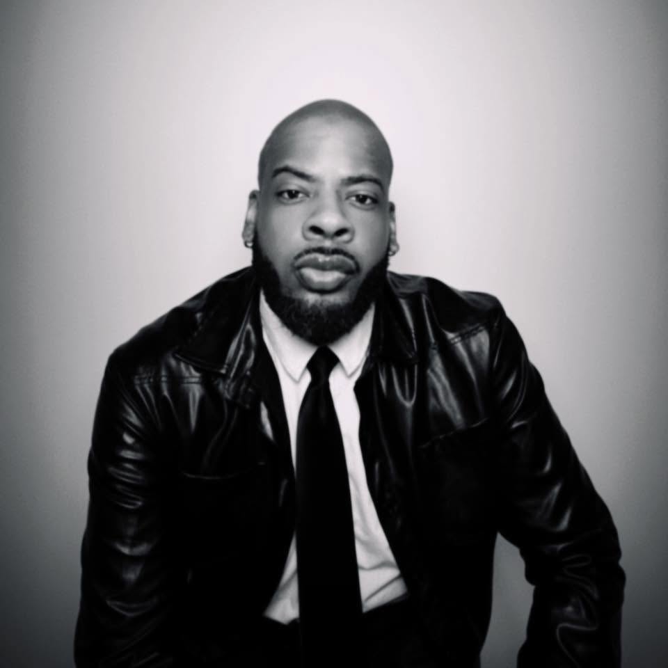 Man with a serious expression, wearing a leather jacket, white shirt, and black tie, against a plain background.