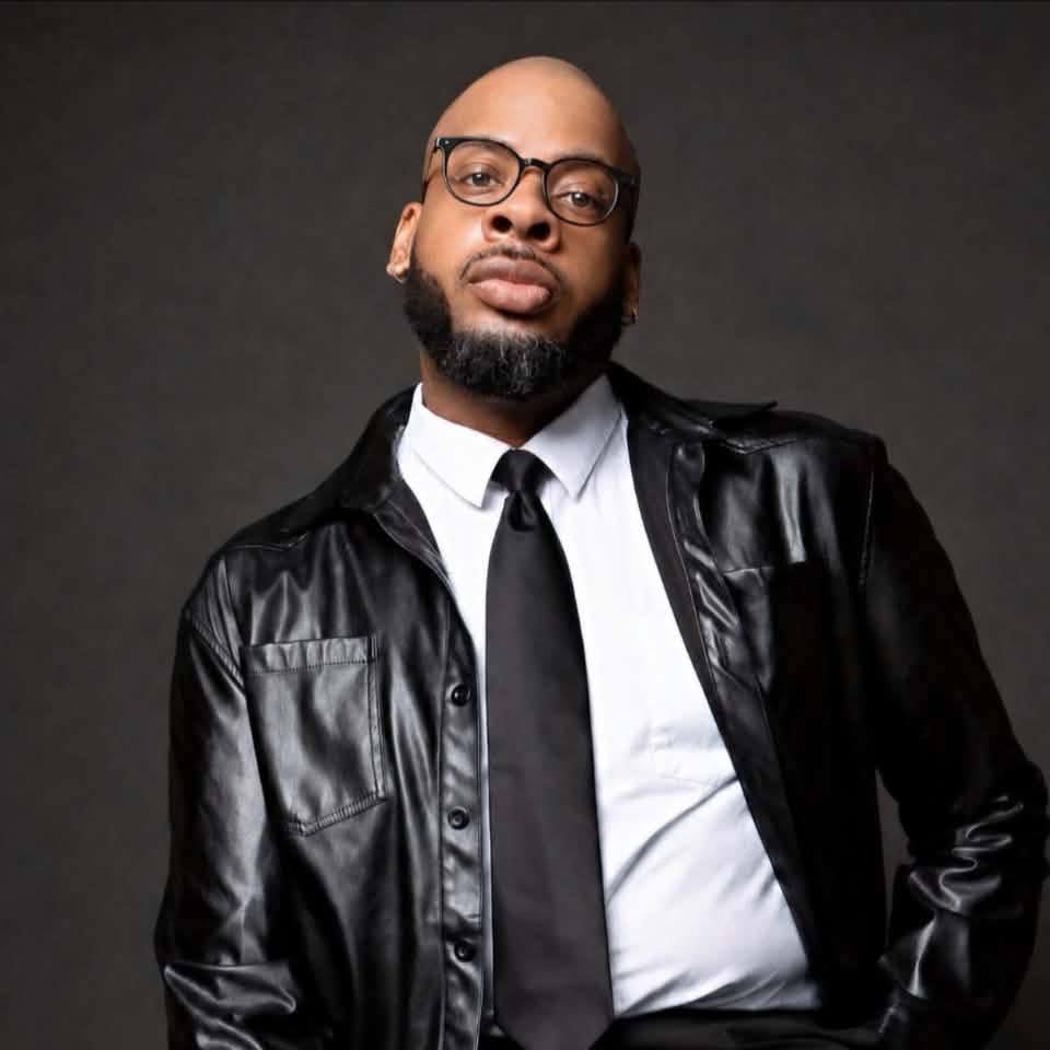 Man with glasses, beard, and bald head wearing a black leather jacket, white shirt, and black tie, posing against a dark background.