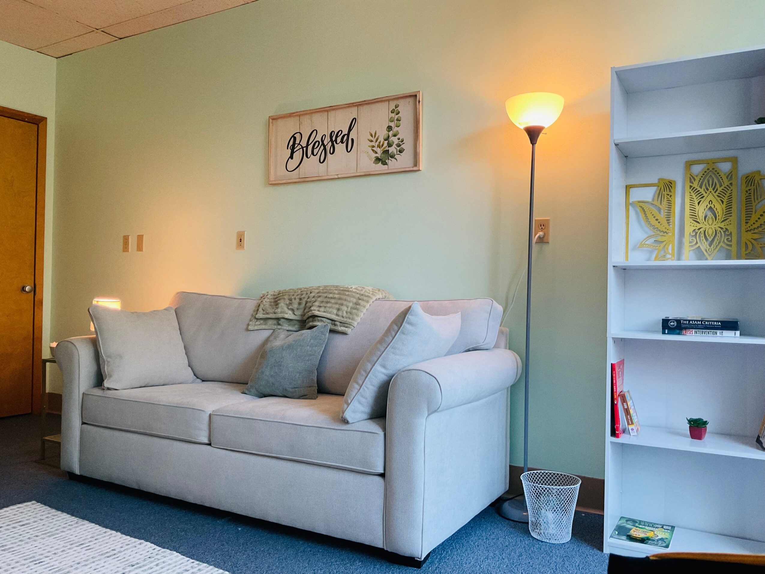 Living room with a beige sofa, wall art, floor lamp, and white bookshelf, against a light green wall.
