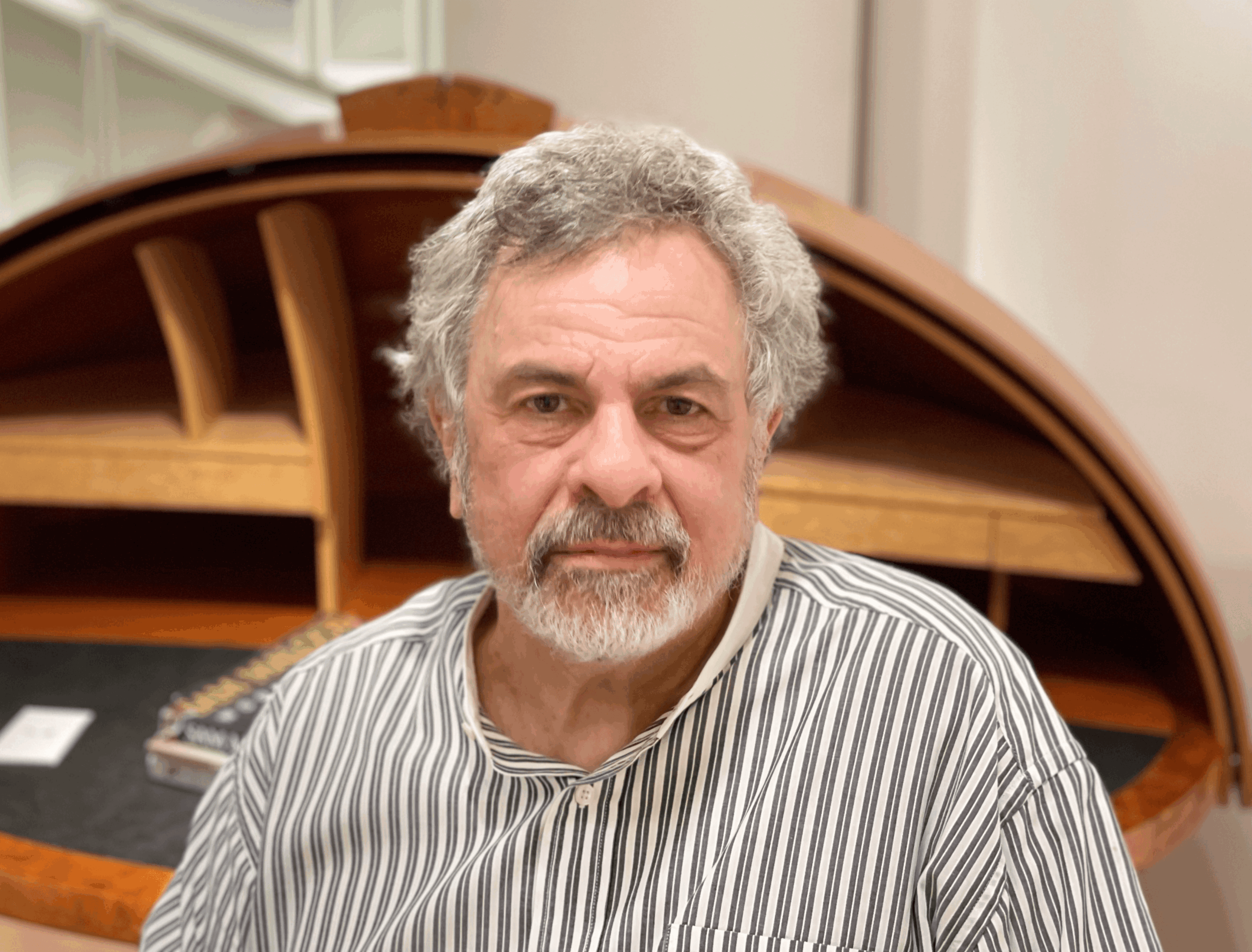 Older man with gray hair and beard, wearing a striped shirt, sitting indoors with a wooden desk in the background.