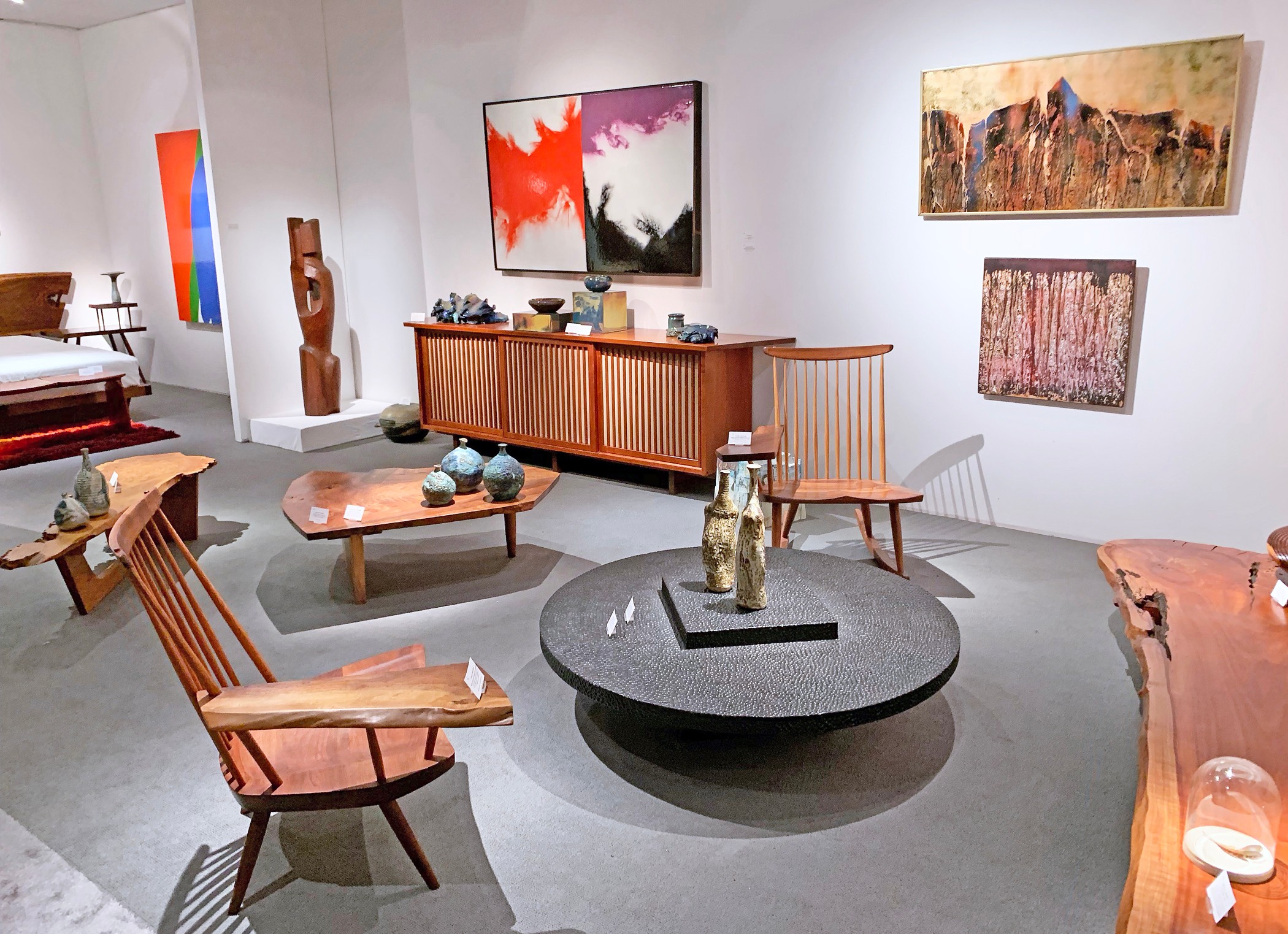 Art gallery with paintings on white walls, wooden furniture, and a round black table with decorative objects.
