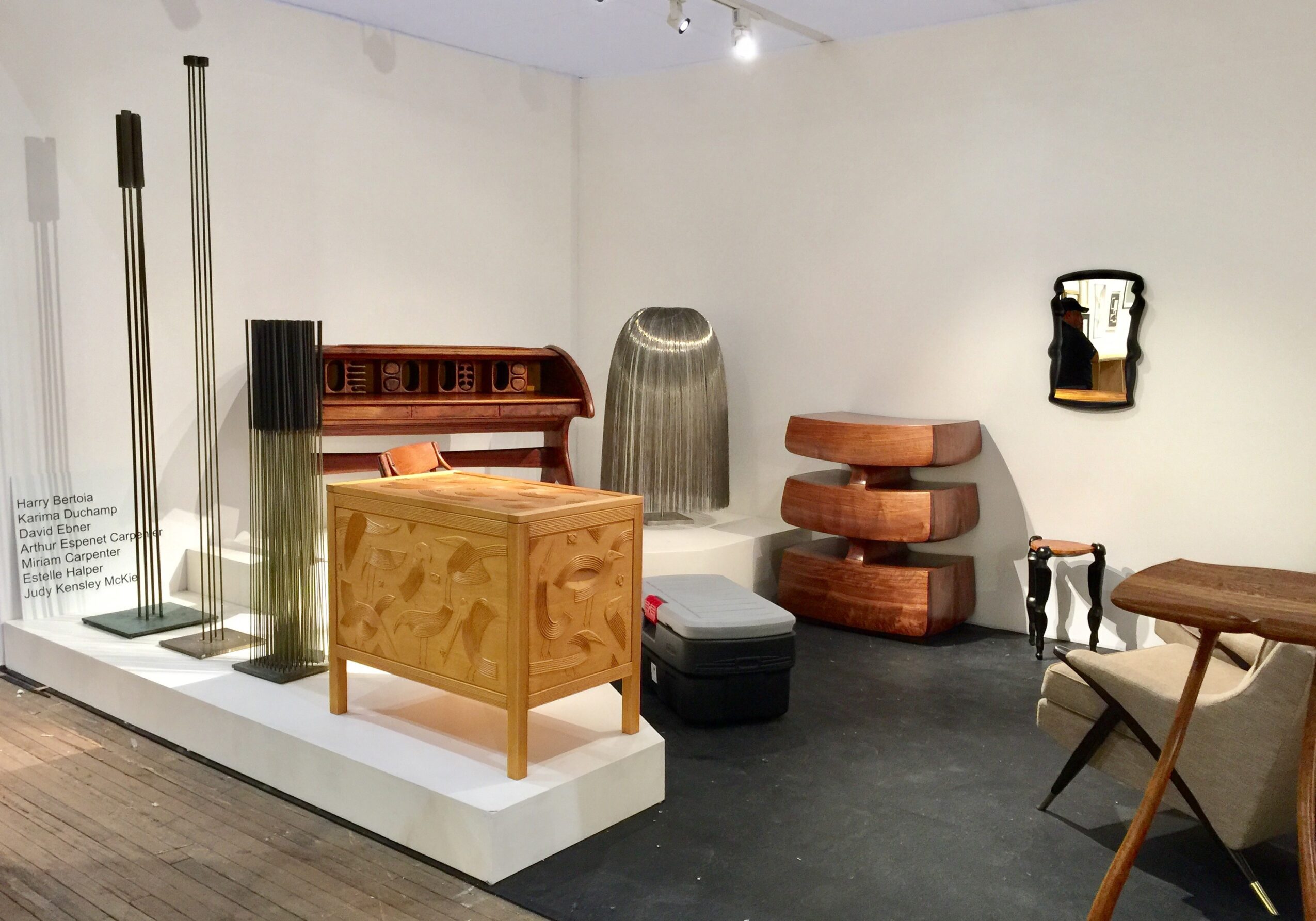 Gallery display with various furniture and decorative objects, including a wooden cabinet, lamps, and chairs, against white walls.