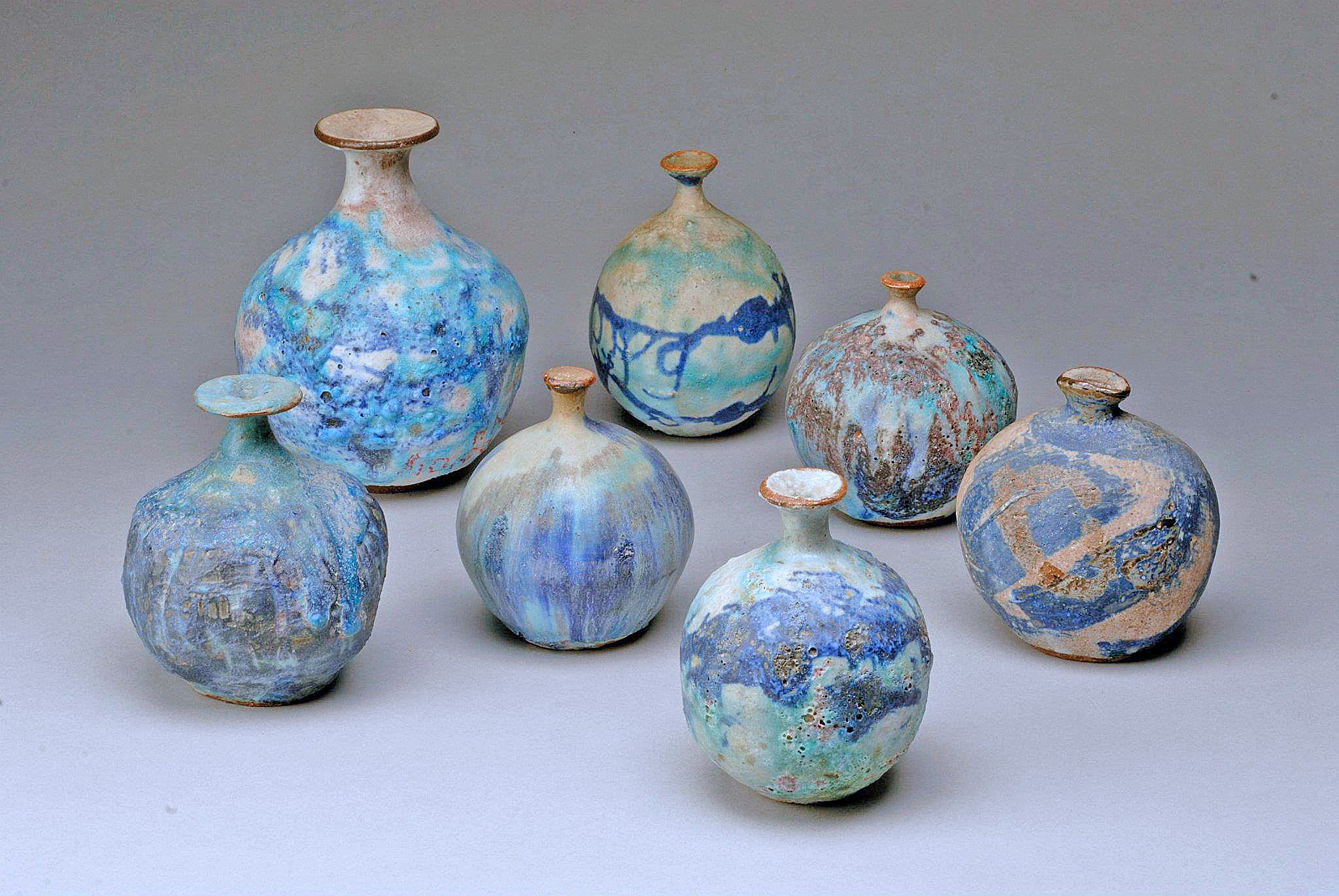 Seven small ceramic vases with blue, green, and brown glazes arranged on a light gray background.