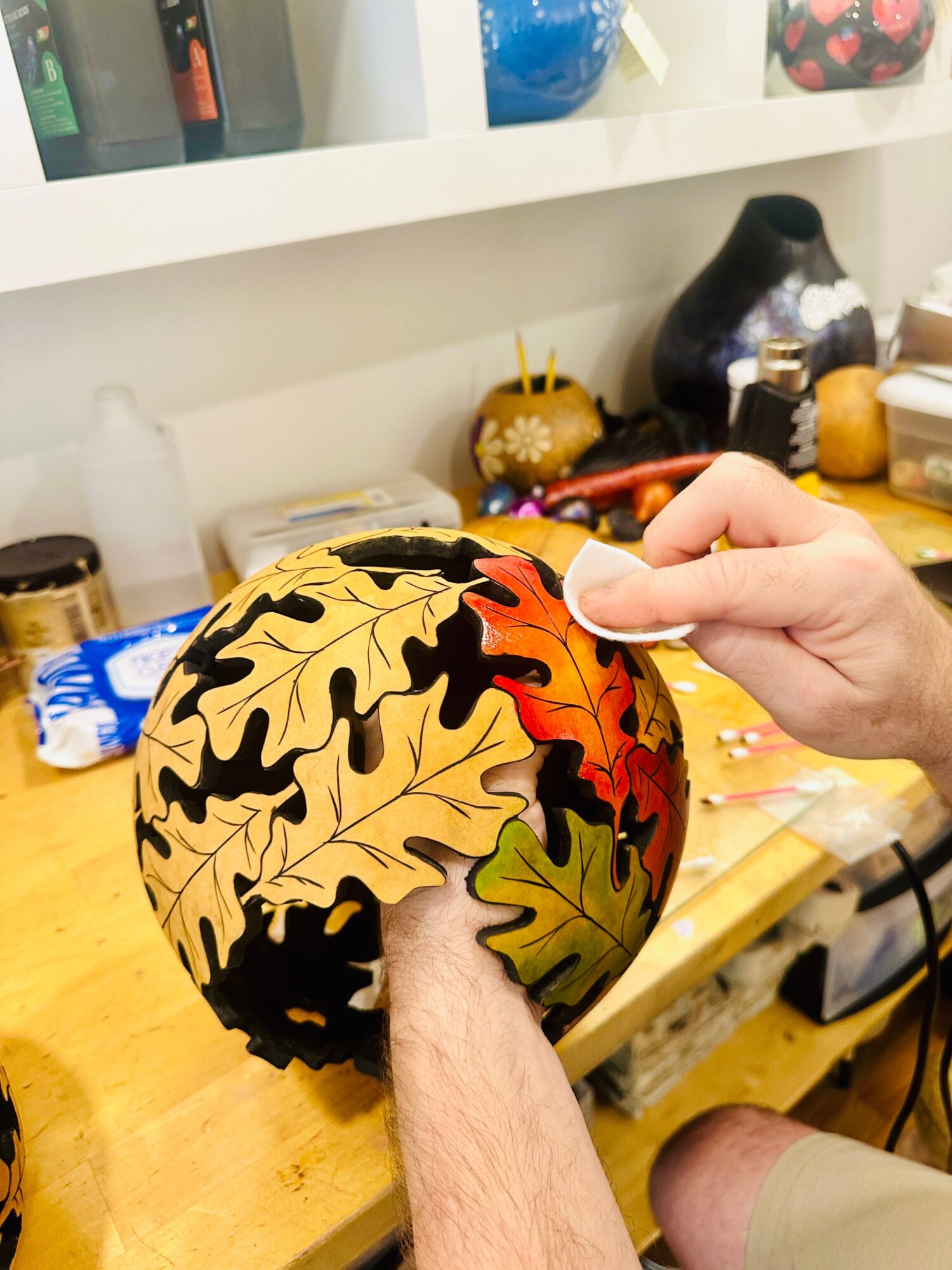 Person holding a decorated pumpkin covered with autumn leaves, in a craft workspace.