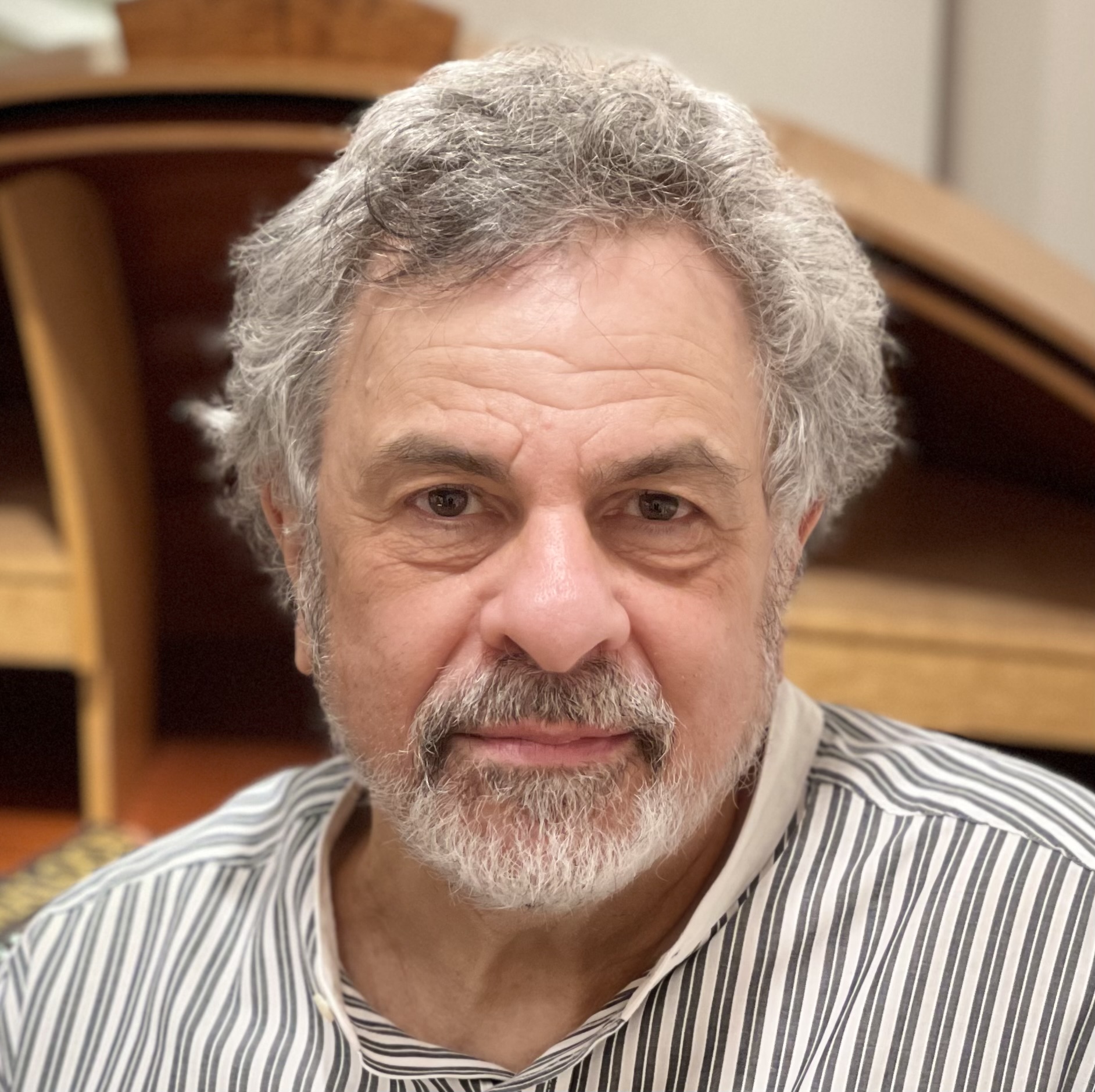 Portrait of an older man with gray hair and beard, wearing a striped shirt, indoors with wooden furniture in background.