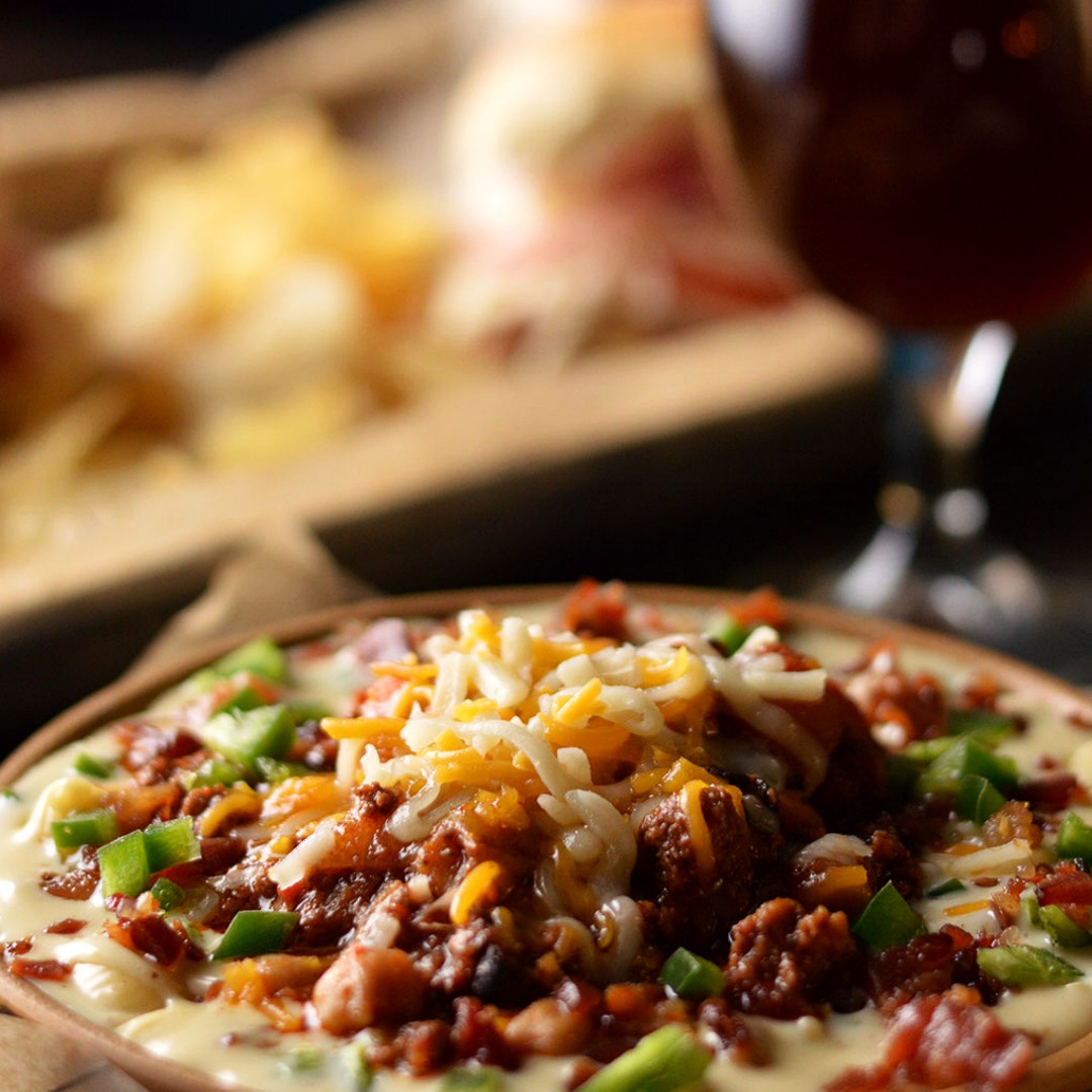 Close-up of a dish with melted cheese, ground meat, and chopped green onions, with a glass of red wine in background.