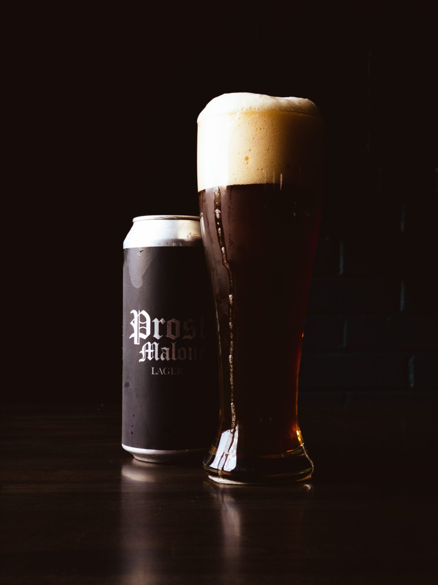 A tall glass of dark beer with foam on top, next to a black can with white text, on a dark surface.
