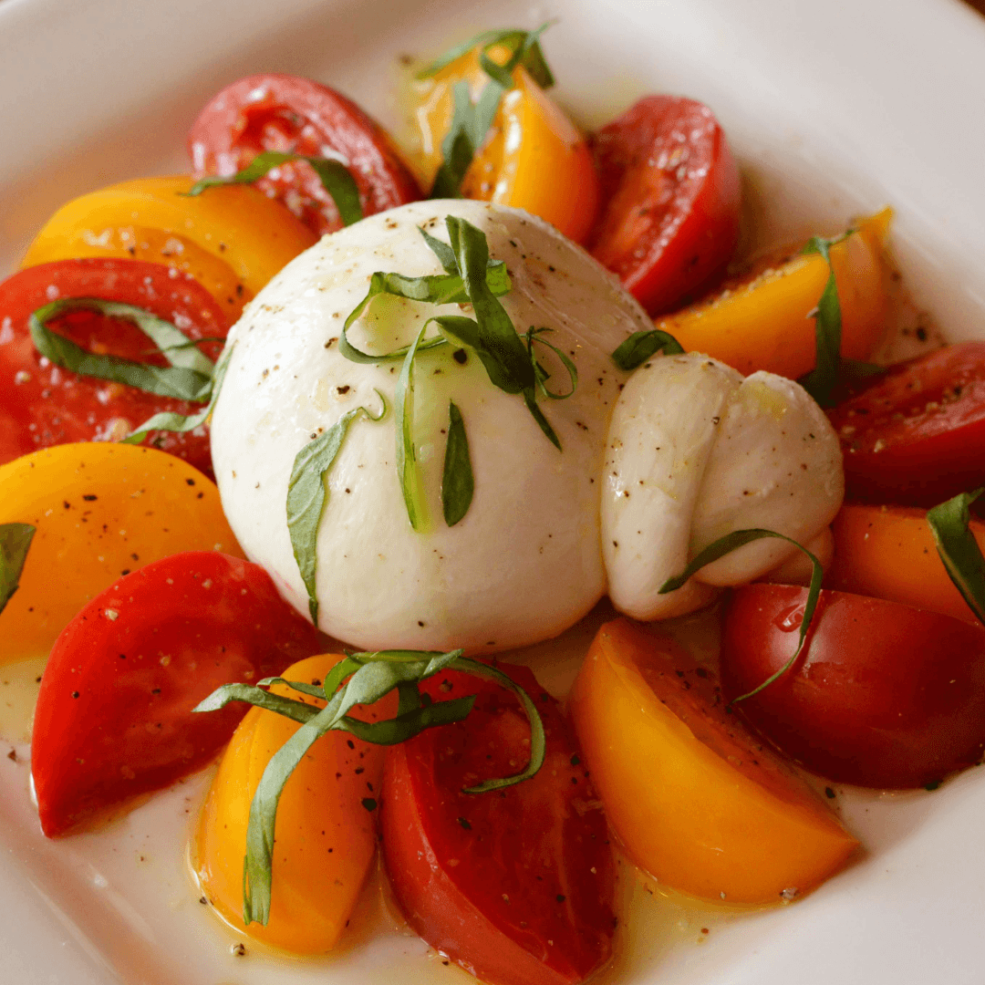 Fresh mozzarella cheese with sliced tomatoes and basil, drizzled with olive oil, on a white plate.
