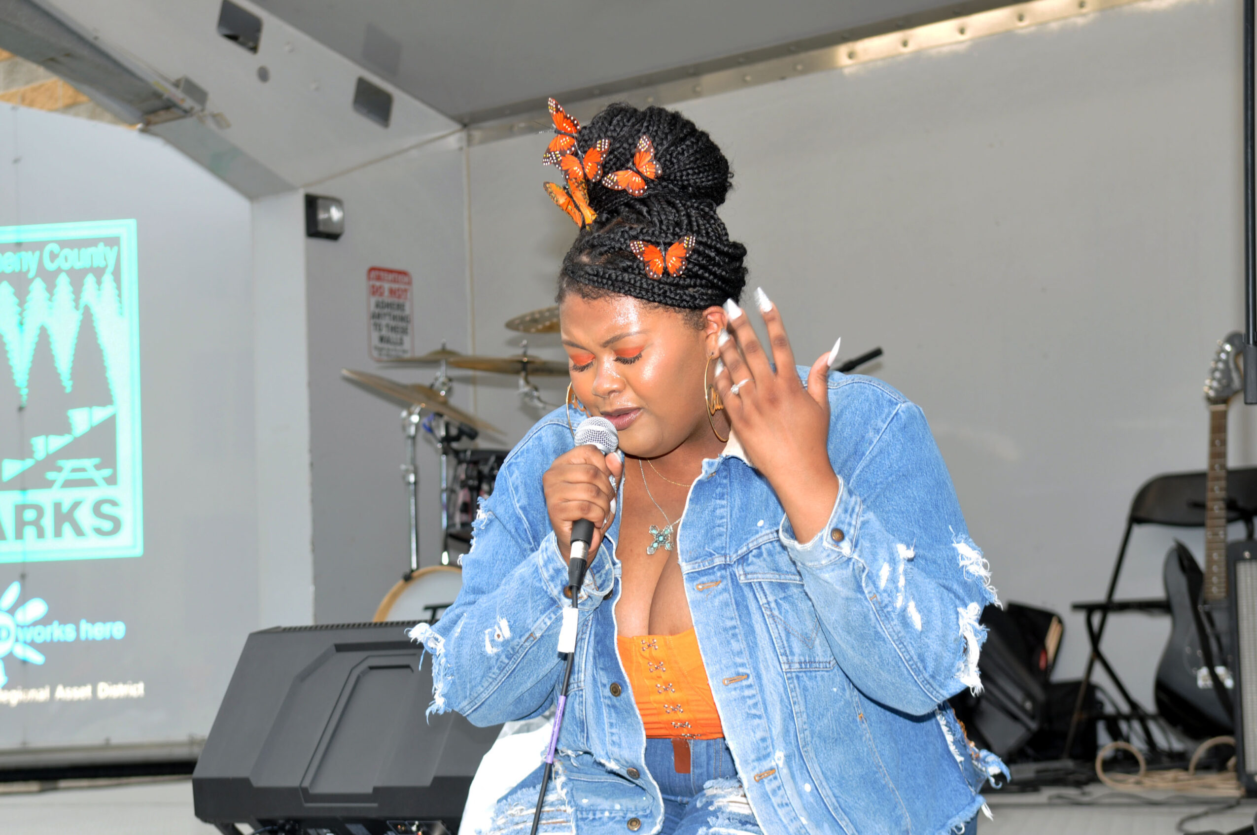 Woman singing into a microphone on stage, wearing a denim jacket and orange top, with a large braided hairstyle decorated with orange accessories.