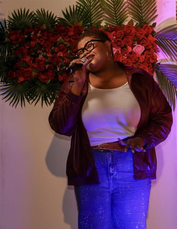 Person singing into a microphone, standing in front of a floral backdrop with red and pink flowers and green leaves.
