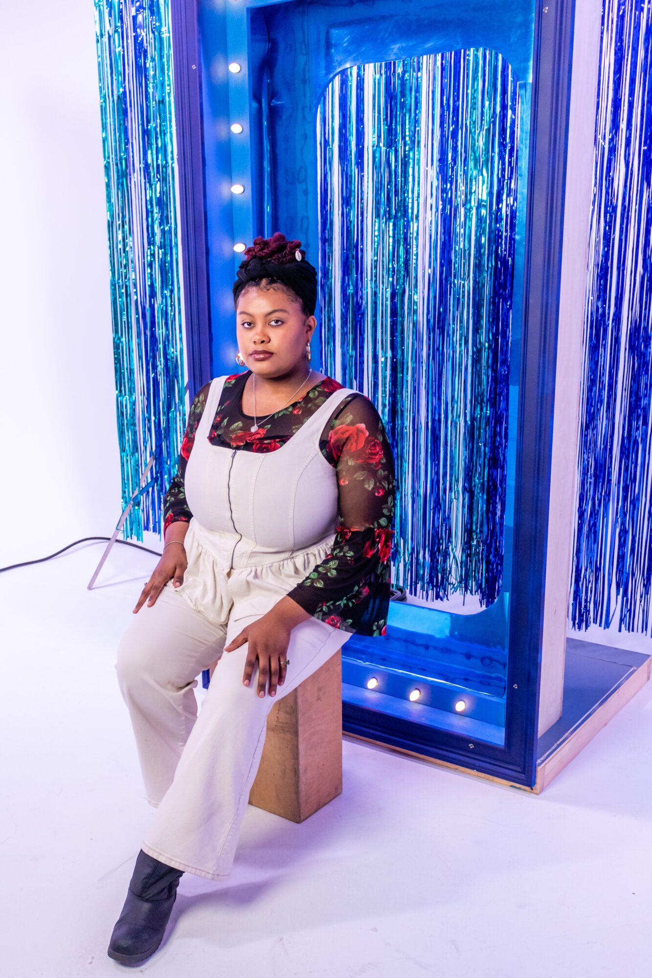 Woman sitting on a wooden block in front of a blue and silver curtain backdrop, wearing a white jumpsuit with black and red lace top.