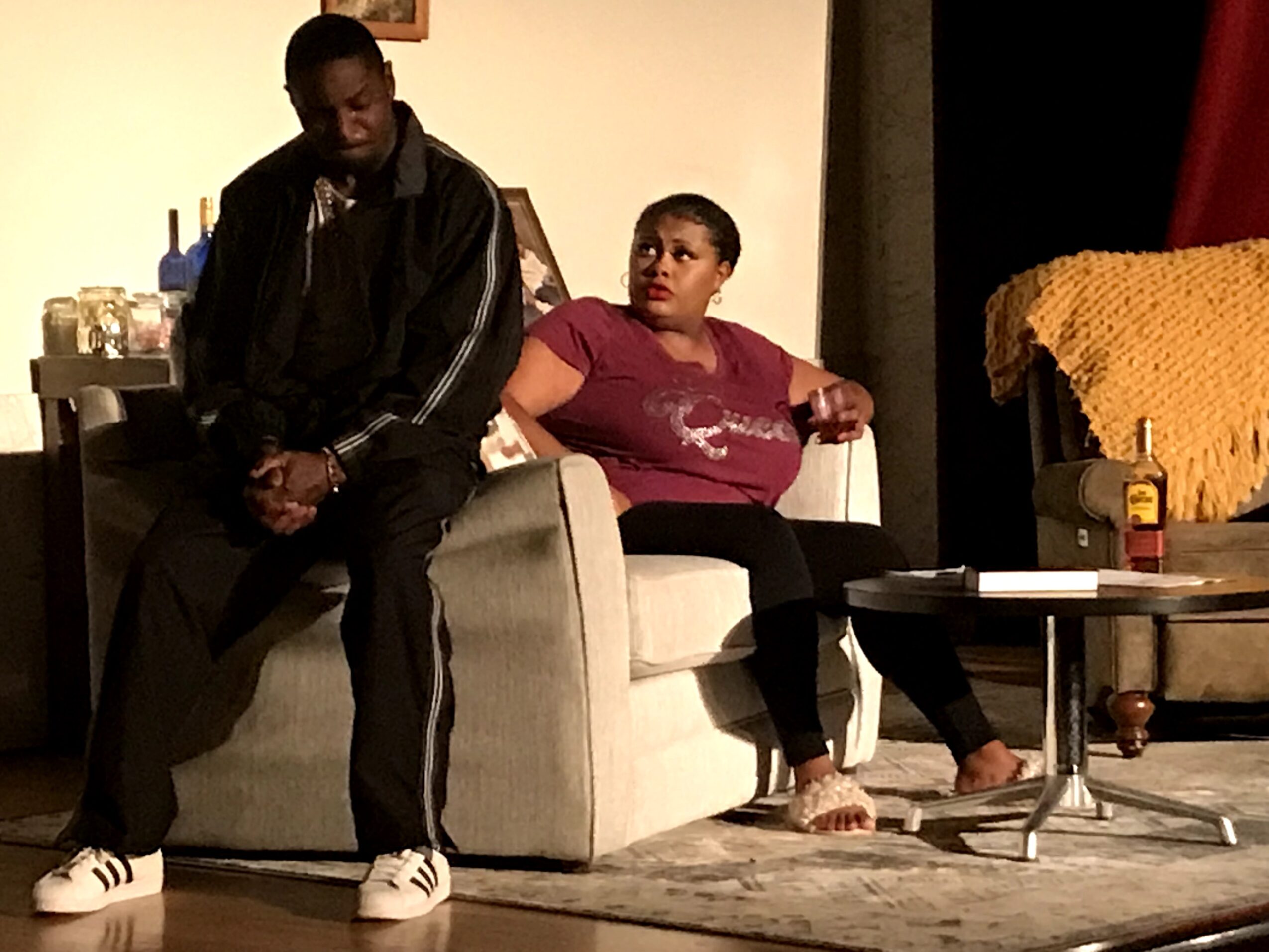 Two people sitting on a sofa in a living room, one person standing nearby, with a table and a blanket in the background.
