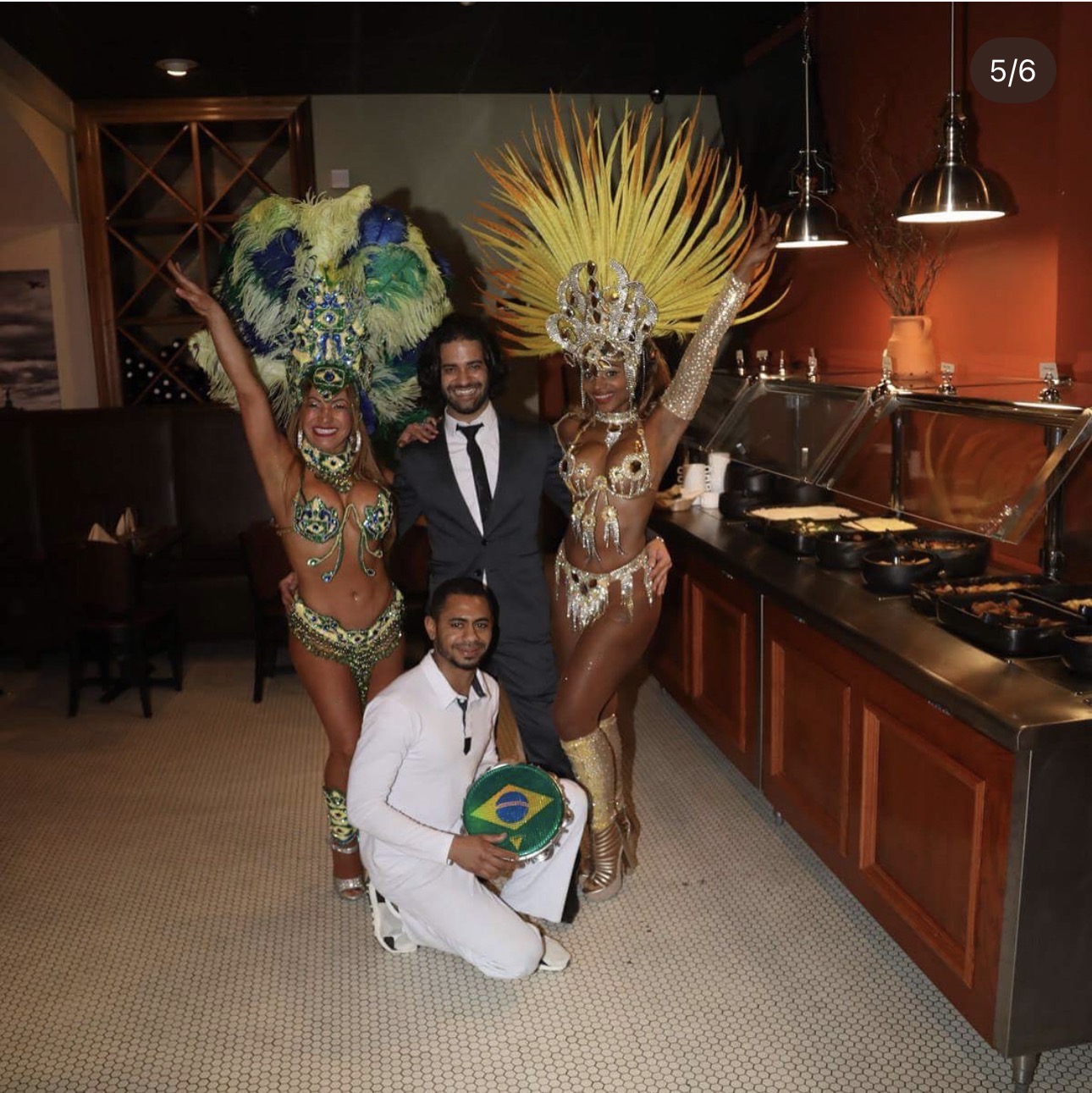 Four people, two in elaborate costumes with feathered headdresses, pose in a restaurant. One person holds a Brazilian flag. All are smiling.