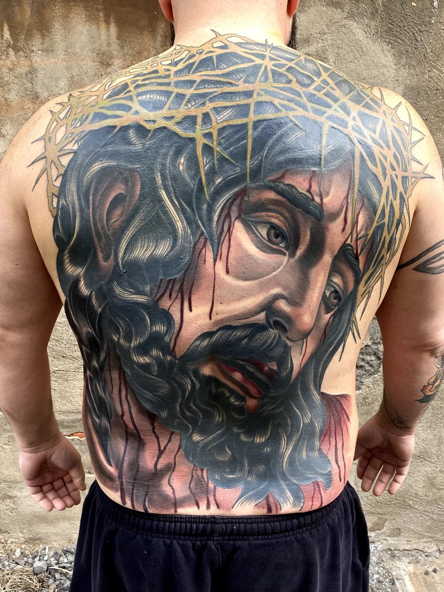 Detailed tattoo of a bearded man's face with a crown of thorns on the back, covering the upper back and shoulders.