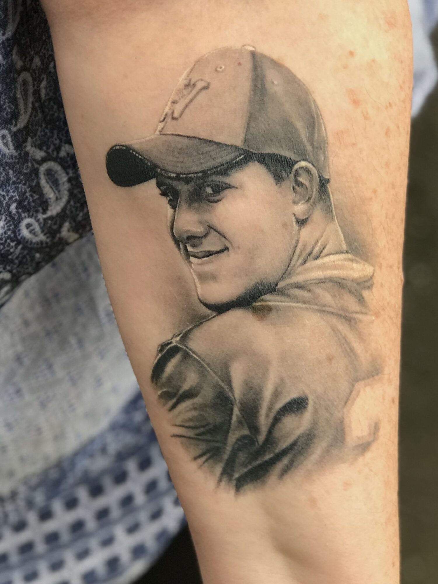 Black and gray tattoo of a smiling person wearing a baseball cap, facing slightly to the right, on skin.