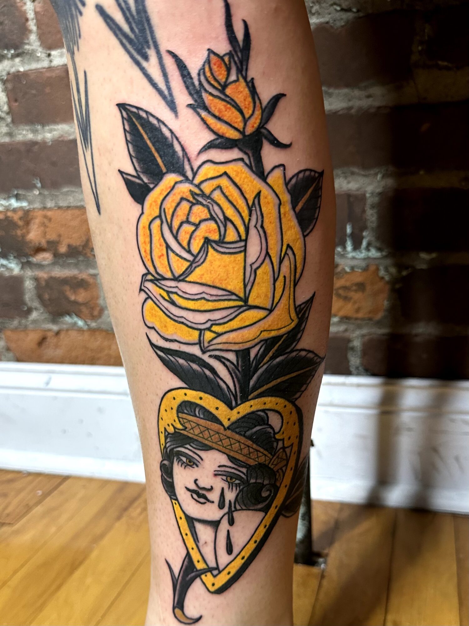 Tattoo of a yellow rose with leaves and a smaller bud, below a woman's face inside a heart-shaped frame, on a person's arm.