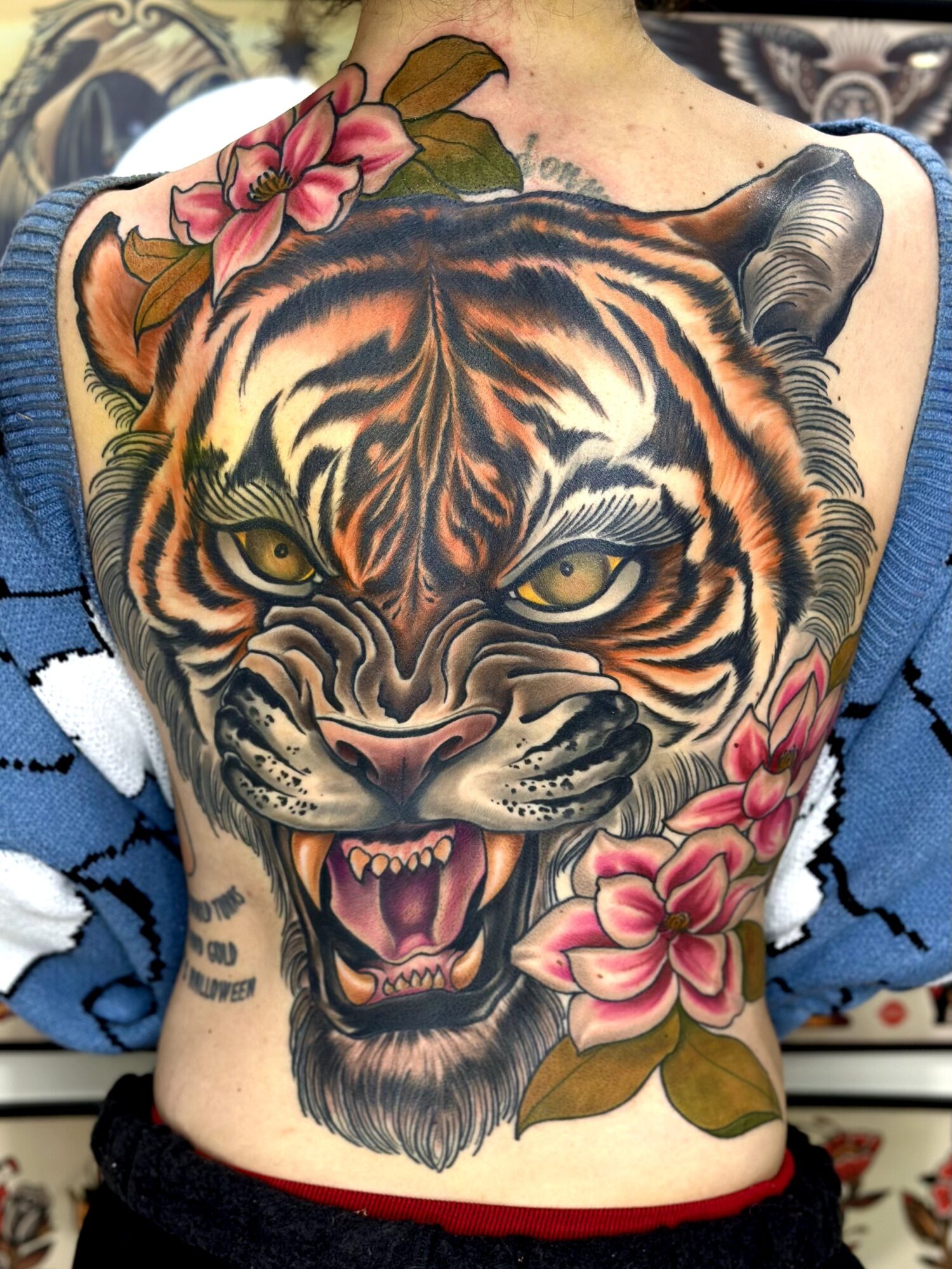 Large tiger face tattoo with open mouth, sharp teeth, and yellow eyes, surrounded by pink flowers and leaves.