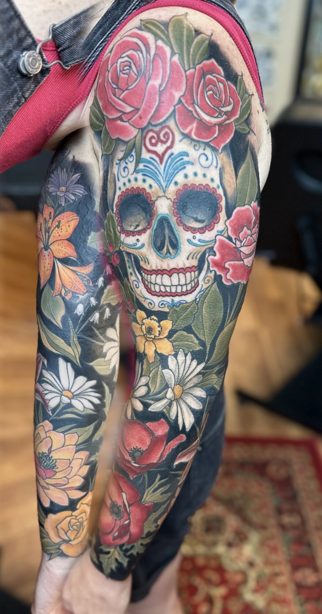 Person with a colorful skull and floral tattoo sleeve on arm and shoulder, featuring roses, daisies, and other flowers.