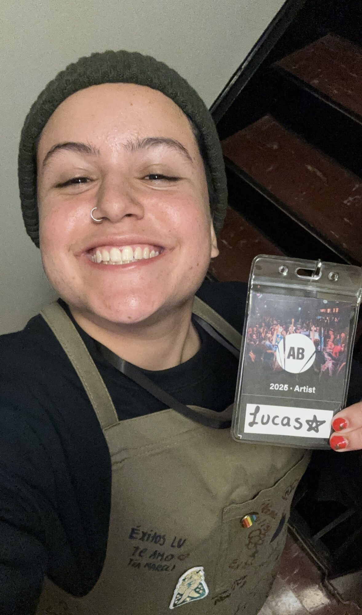 Person smiling, wearing a beanie and apron, holding a badge with the name 'Lucass' and a star, in front of stairs.