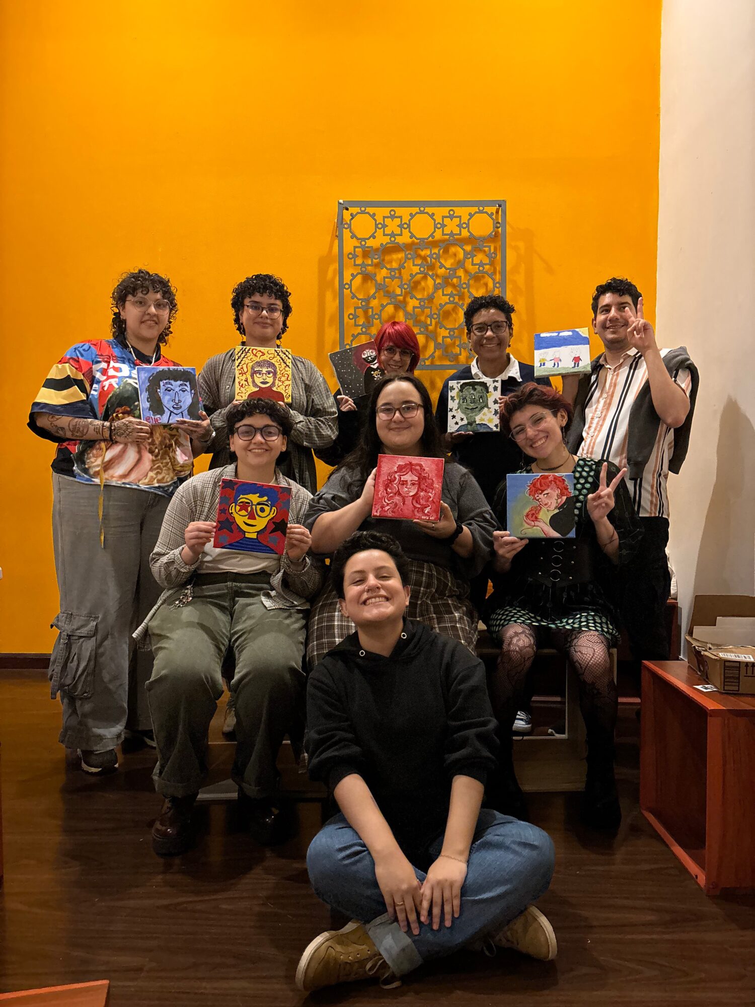 Group of nine people, some holding colorful artwork, posing indoors against a yellow wall with a decorative window, smiling.