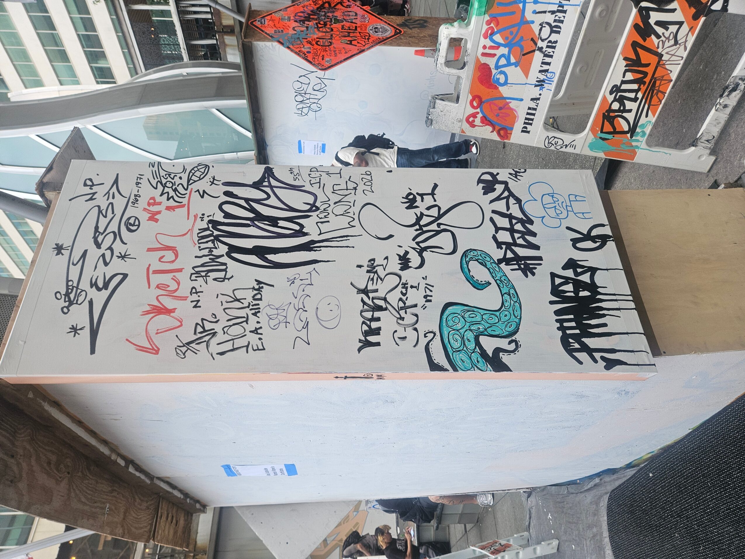 Vertical white surface covered with various graffiti and signatures, with a city street and buildings in the background.