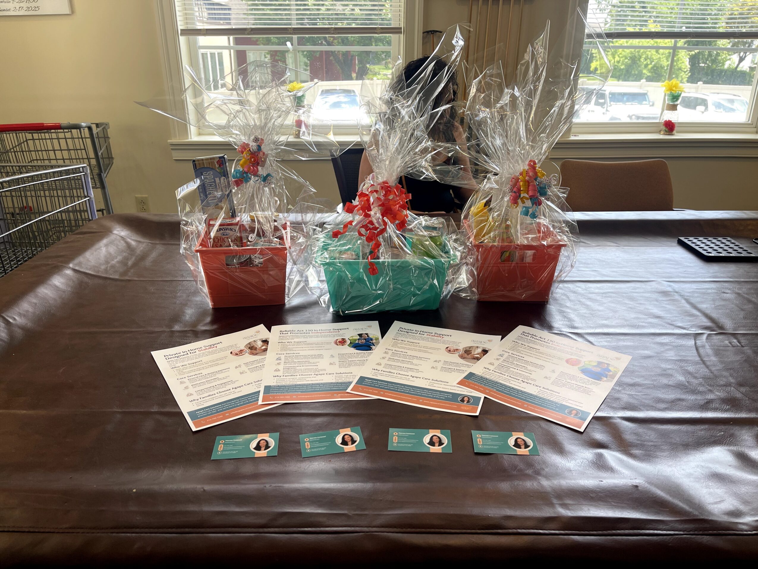 Three gift baskets wrapped in clear plastic with ribbons, placed on a table with documents and cards in front, in a room with windows.