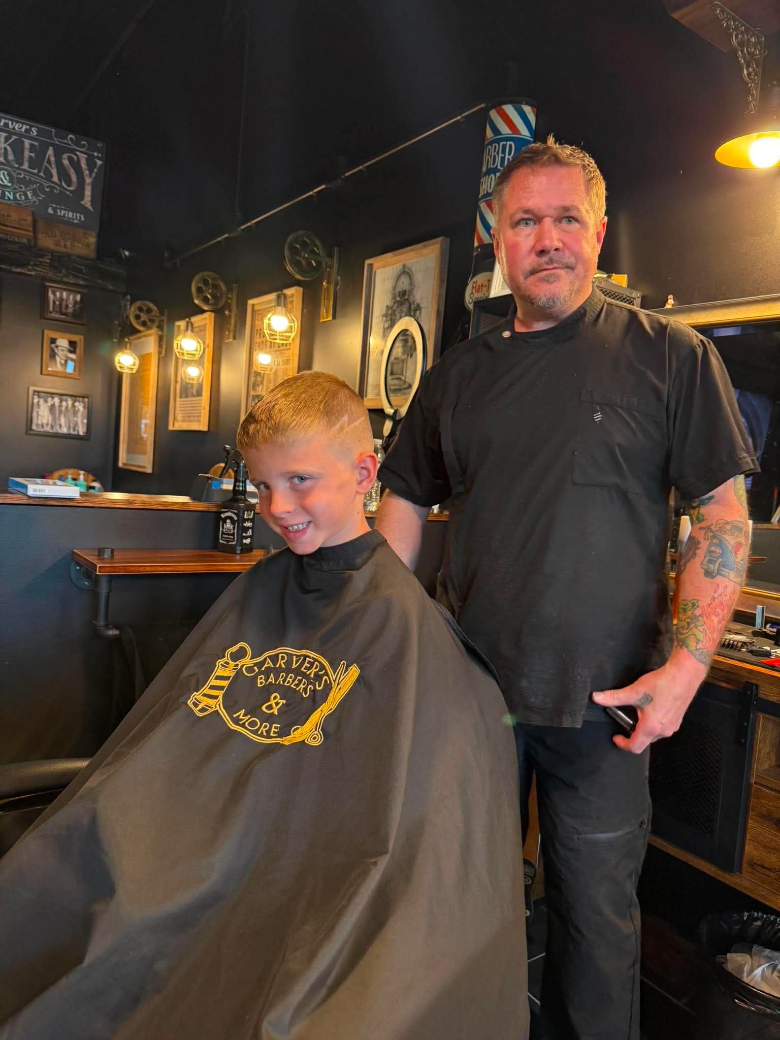 A young boy with a smile sits in a barber chair, with a barber standing beside him in a barbershop.